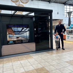 Glass Area - Nowoczesna ekspozycja zabudowy tarasu z czarną ramą w Jastrzębnikach. Na ekspozycji widoczny plakat z wizualizacją szklanego tarasu. Obok stoi mężczyzna z dzieckiem.