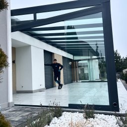 Glass Area - Nowoczesna, przeszklona konstrukcja z czarną ramą, prawdopodobnie zadaszenie tarasu lub weranda. Osoba opiera się o ścianę. Białe kamienie dekoracyjne na pierwszym planie.