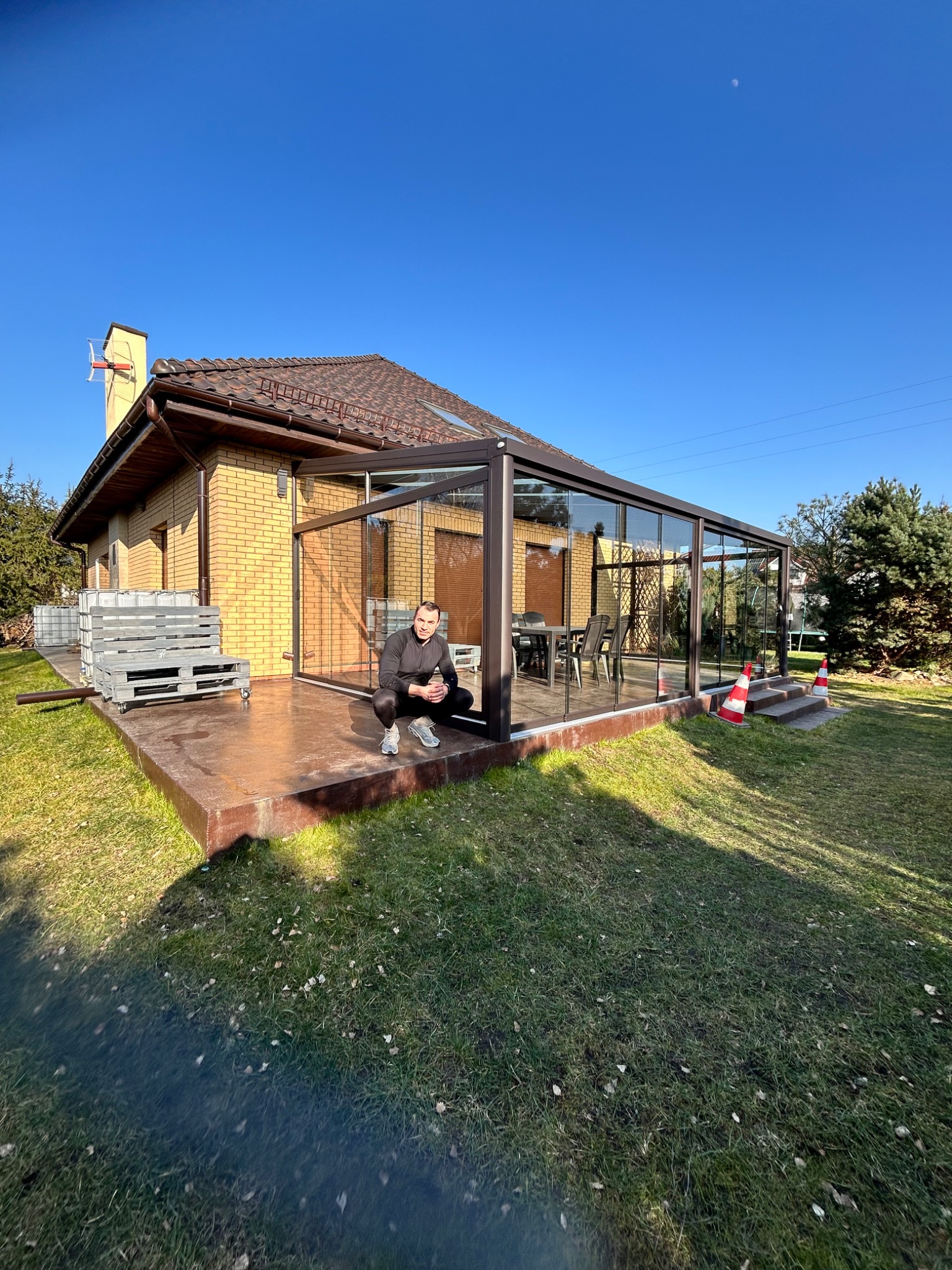 Szklana zabudowa tarasu z brązowymi ramami, zewnętrzna część domu z cegły, siedzący mężczyzna, błękitne niebo. Widok z zewnątrz na gotową konstrukcję.