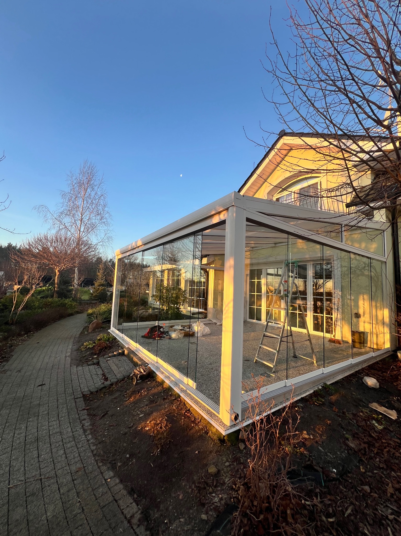 Szklana zabudowa tarasu z białymi profilami w ogrodzie, widok z zewnątrz. Drabina oparta o ścianę, błękitne niebo w tle.