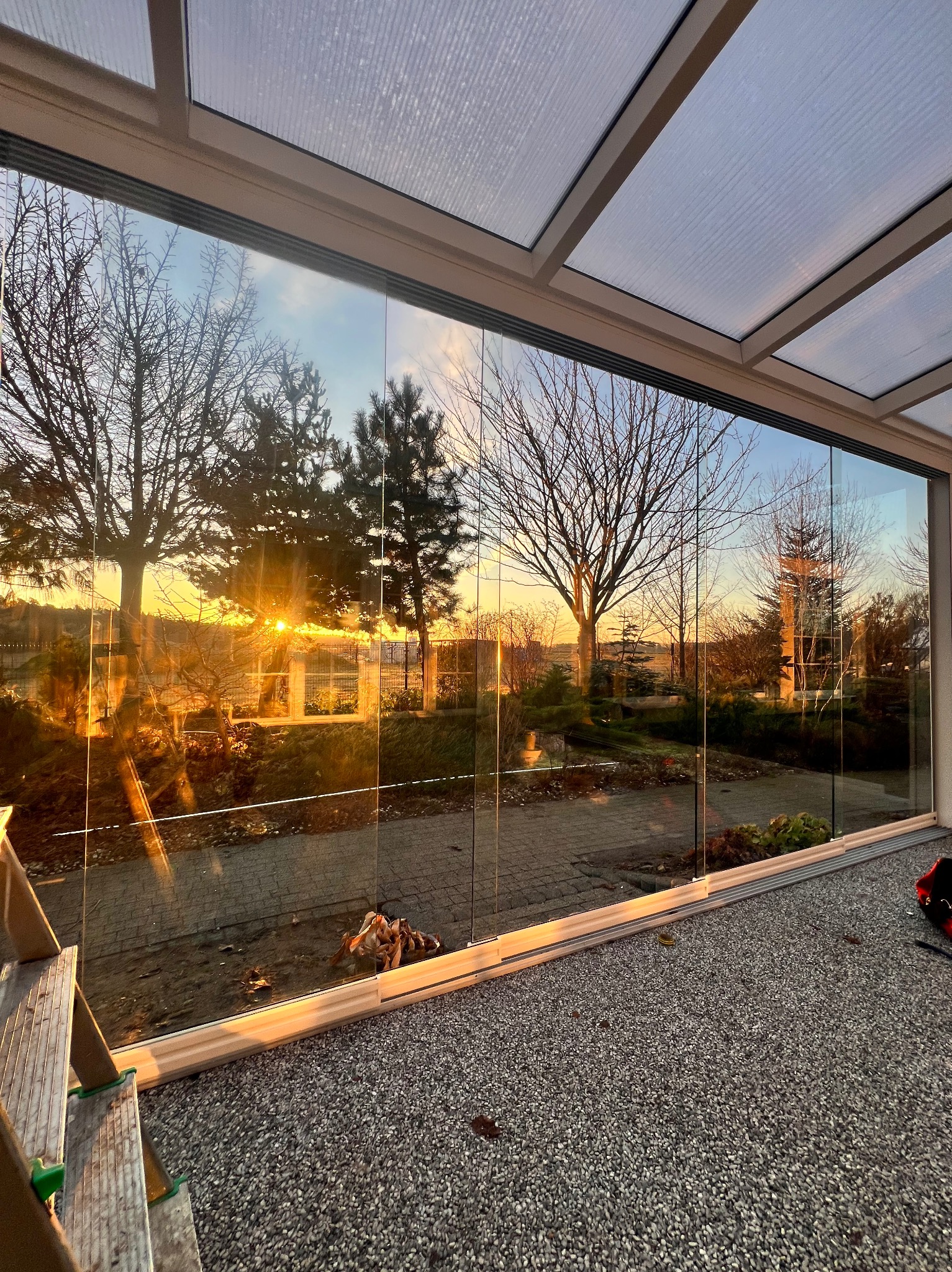 Szklana zabudowa balkonu z widokiem na ogród o zachodzie słońca. Widoczne przesuwne drzwi szklane i metalowa konstrukcja zadaszenia. Częściowo widoczna drabina.