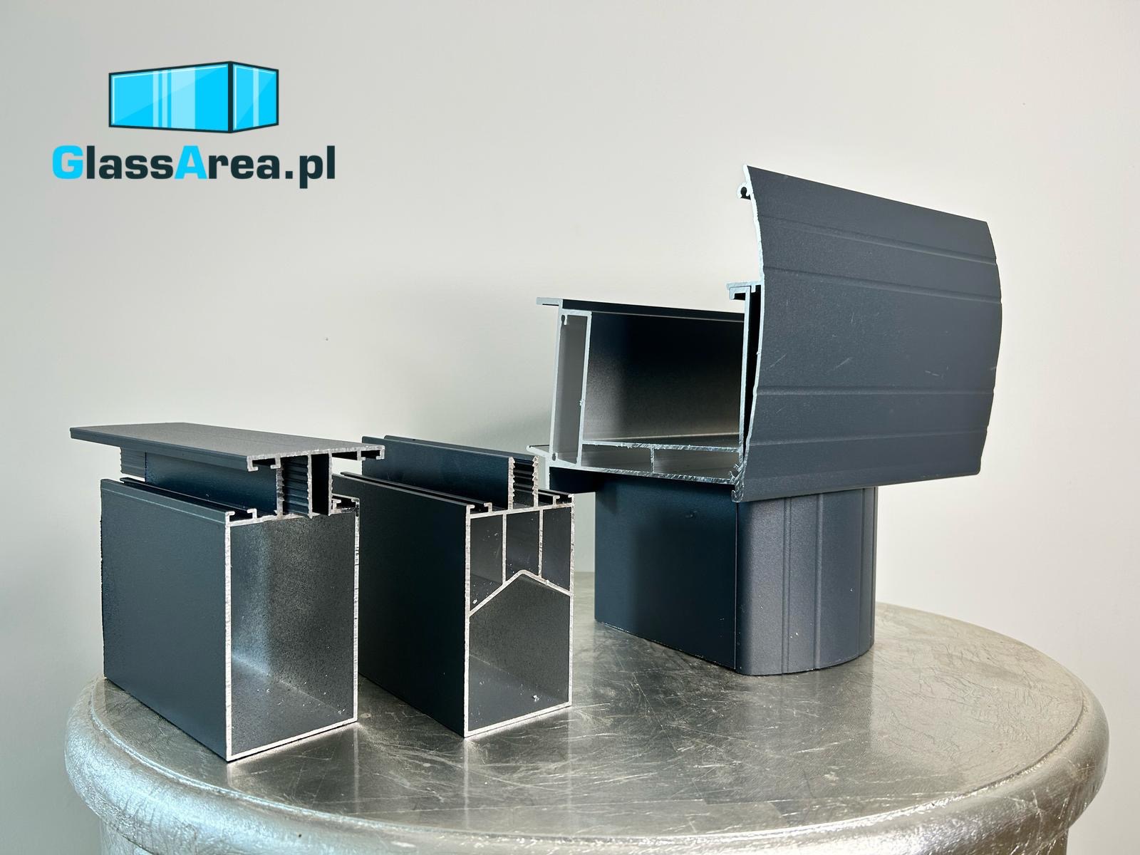 Prezentacja przekrojów profili aluminiowych w kolorze grafitowym na metalowym stole. Widoczne detale konstrukcyjne i wykończeniowe. GlassArea.pl w tle.