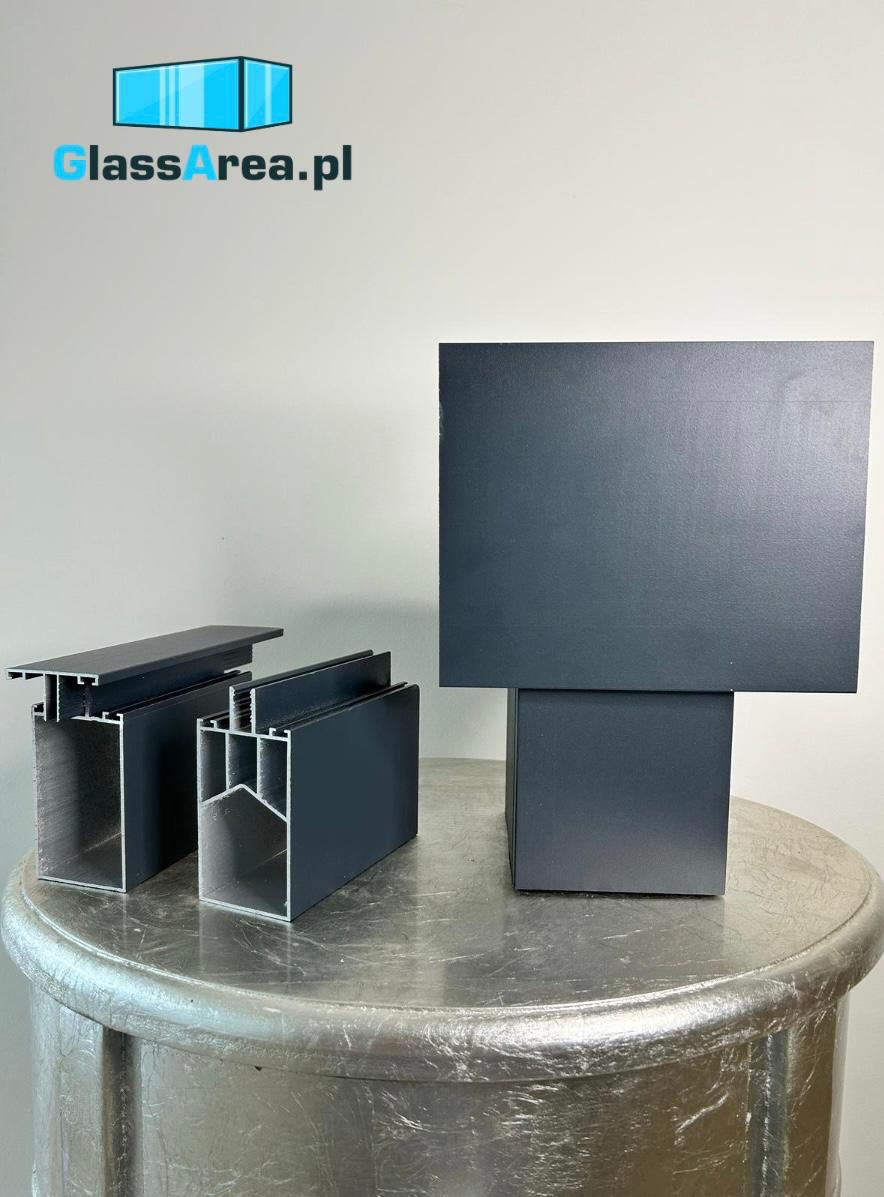 Czarne profile aluminiowe na srebrnym stoliku, prezentujące przekroje i kształty elementów konstrukcyjnych, z logotypem glassarea.pl w tle.