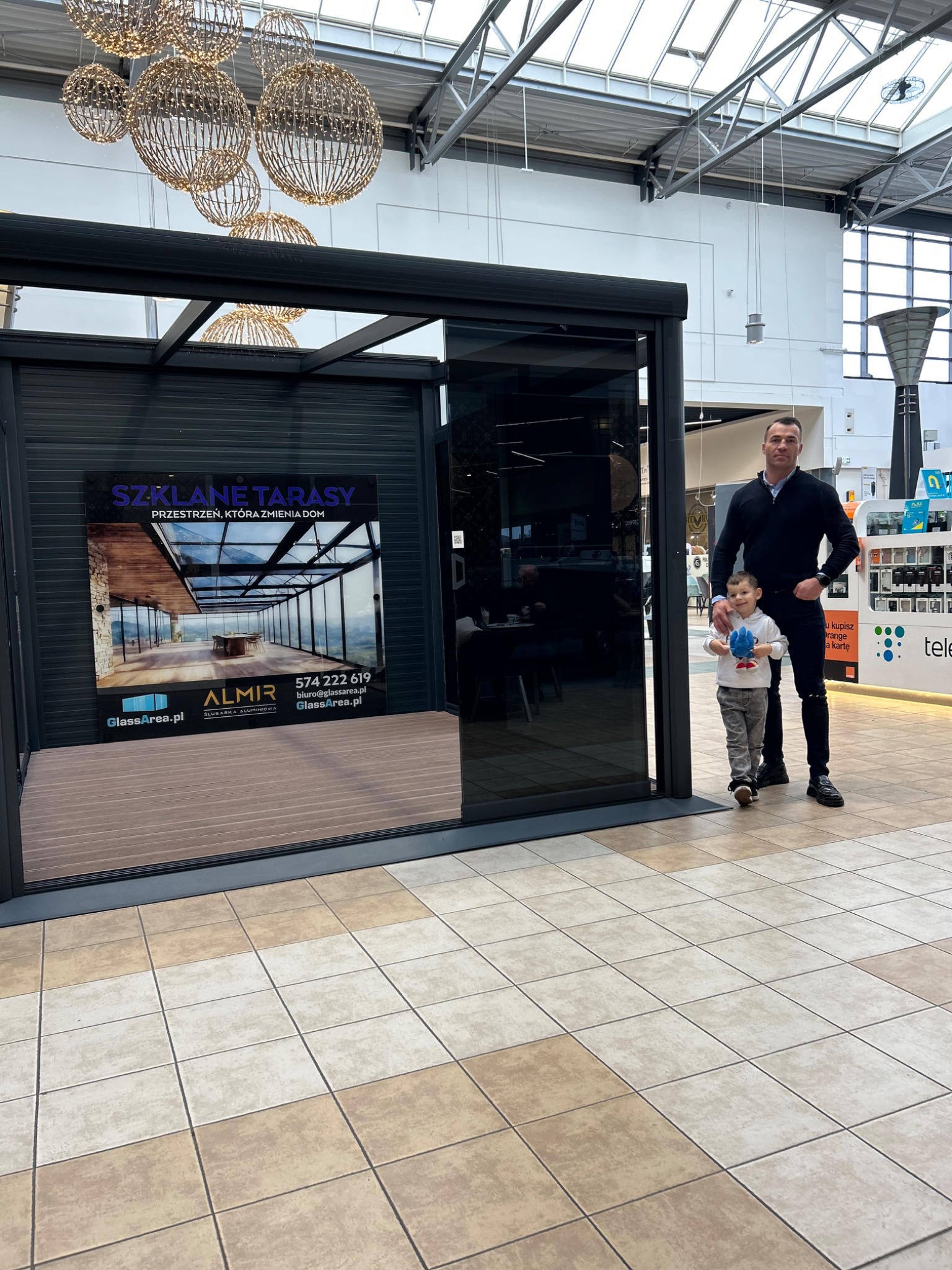Nowoczesna ekspozycja zabudowy tarasu z czarną ramą w Jastrzębnikach. Na ekspozycji widoczny plakat z wizualizacją szklanego tarasu. Obok stoi mężczyzna z dzieckiem.