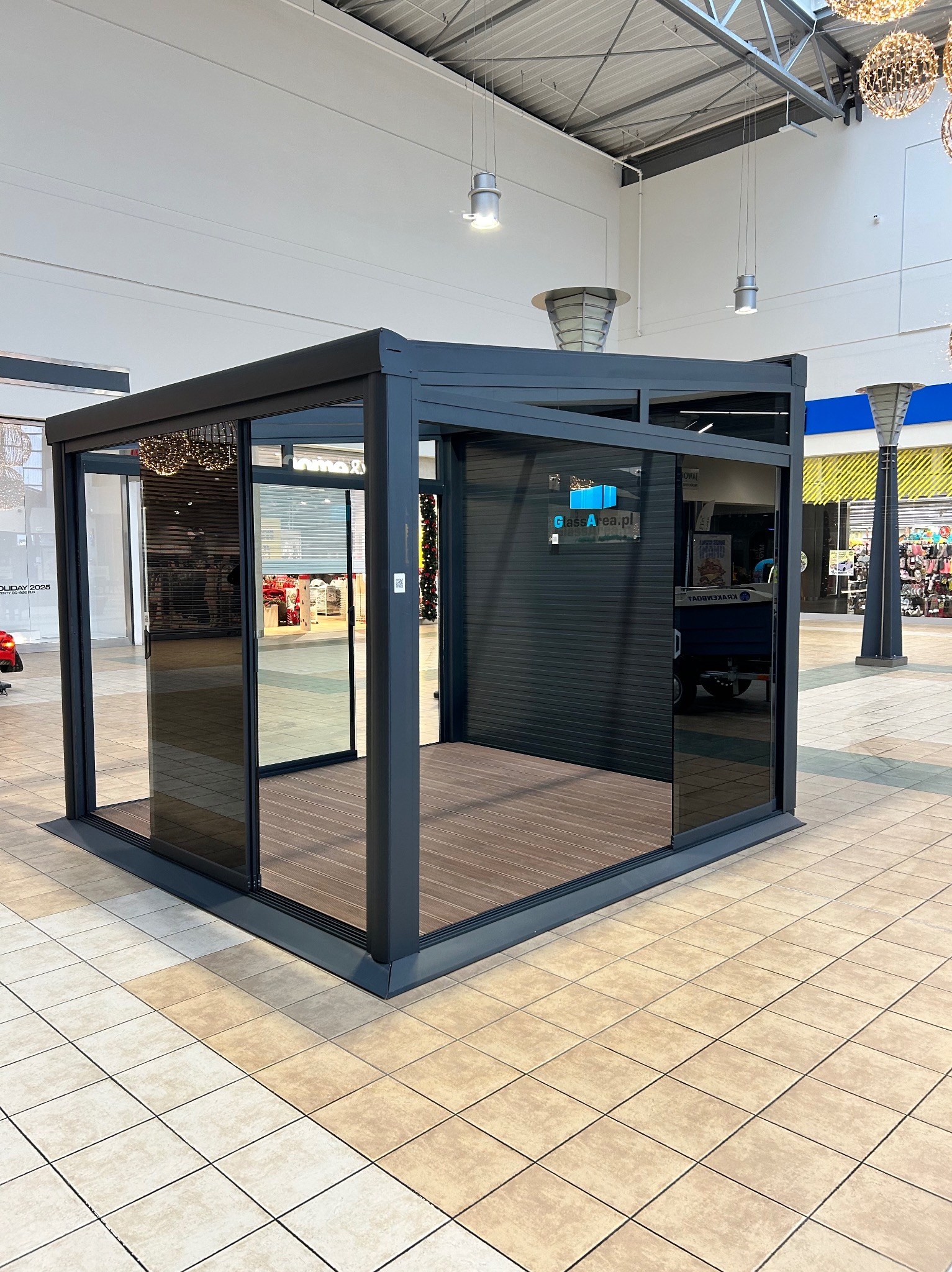 Nowoczesna, przeszklona pergola z ciemną ramą i drewnianą podłogą, umieszczona wewnątrz galerii handlowej. Widoczne przesuwne drzwi i logo firmy na szybie.