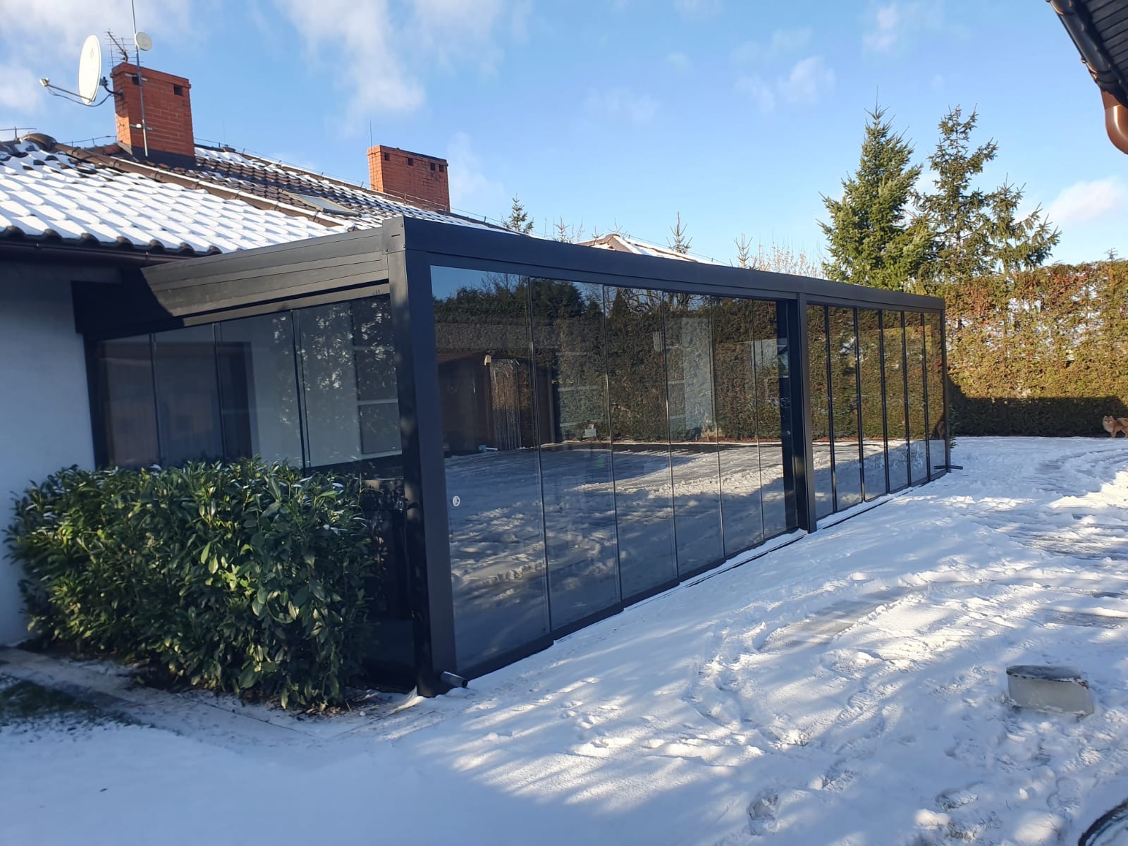 Zimowa zabudowa tarasu z czarną ramą i szklanymi ścianami, odbijająca otoczenie. Śnieg na ziemi i dachu domu. Krzew przed konstrukcją.