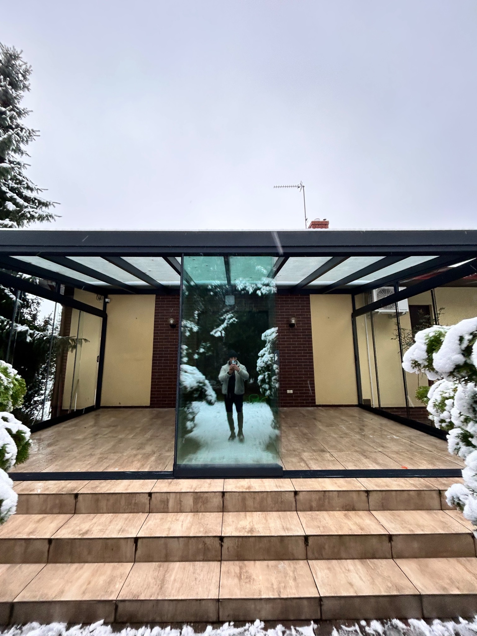 Zadaszony taras z metalową konstrukcją i szklanymi ścianami w zimowej scenerii. Widoczne schody wejściowe i odbicie osoby robiącej zdjęcie w szklanej tafli.