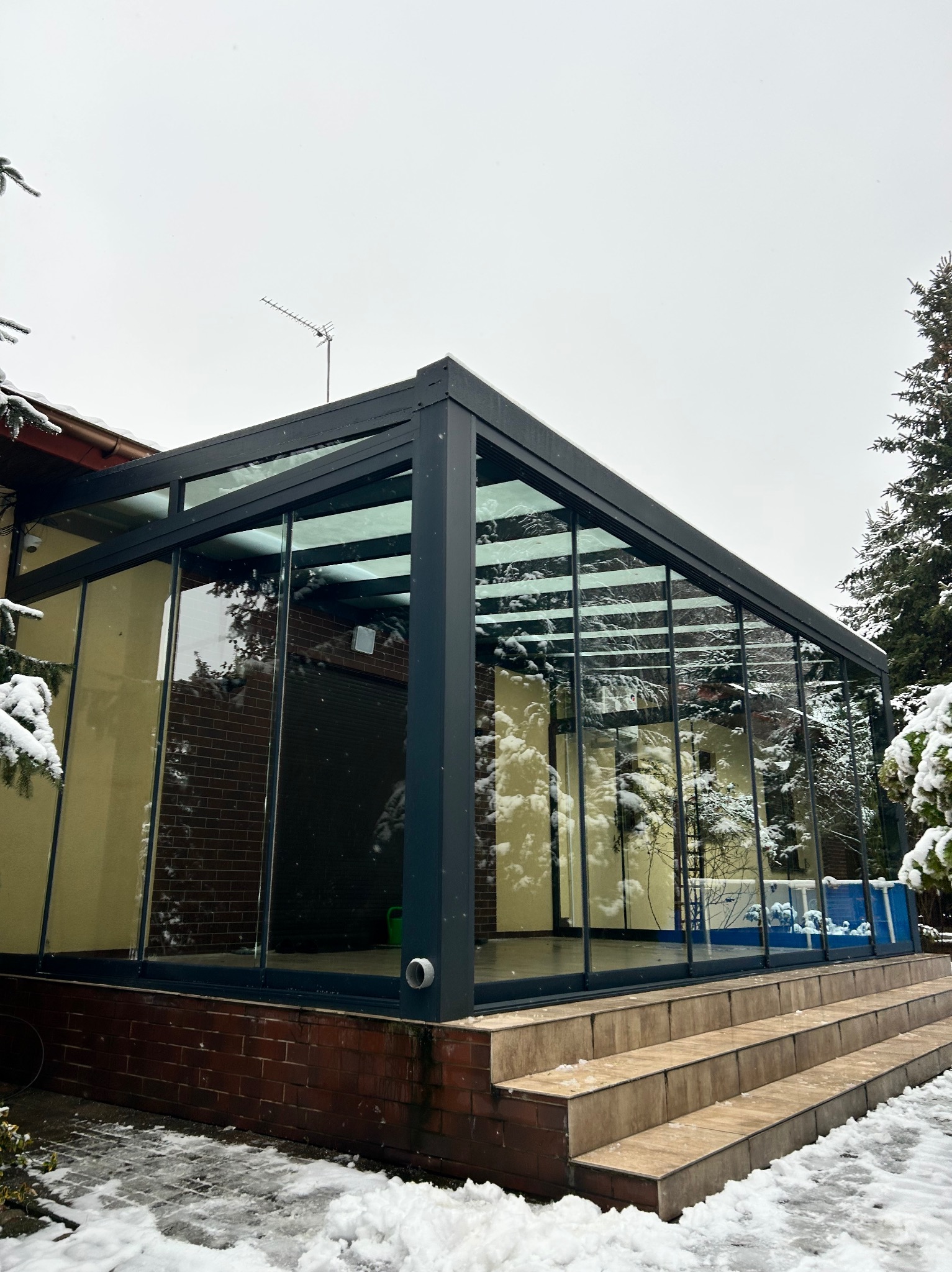 Zimowa zabudowa tarasu z czarną ramą i szklanymi ścianami, widok z zewnątrz na schody wejściowe pokryte śniegiem. Na dachu antena.