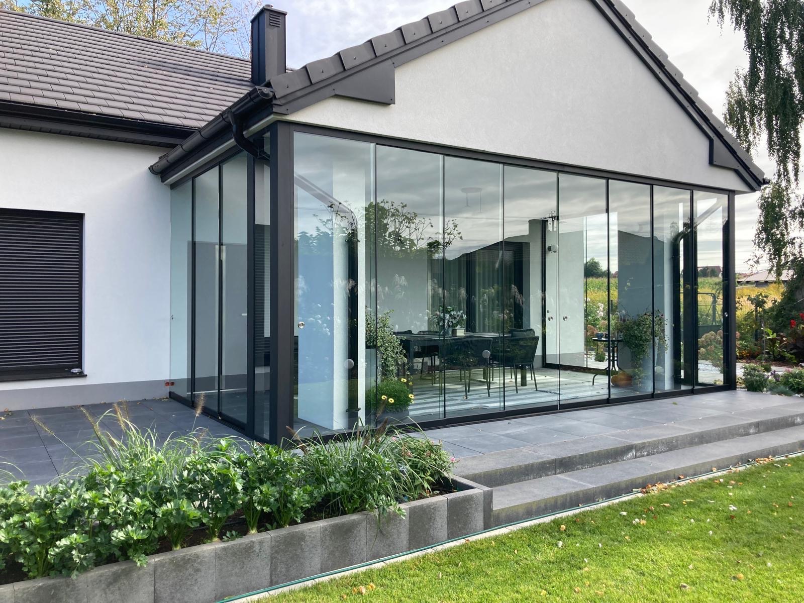 Nowoczesna, przeszklona zabudowa tarasu z czarnymi ramami, widok z zewnątrz na eleganckie wnętrze. Minimalistyczny design, rośliny w donicach, kamienne stopnie.