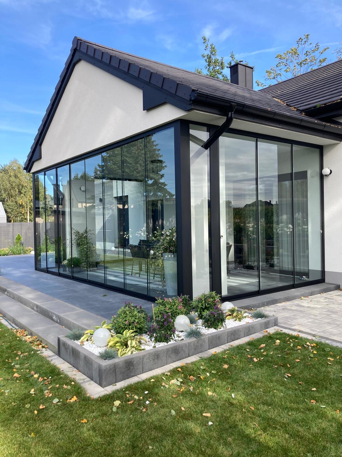 Nowoczesna, przeszklona zabudowa tarasu z czarnymi ramami, widok z zewnątrz. Uwagę przykuwa starannie zagospodarowany ogródek przed zabudową.