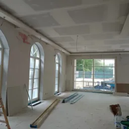 Wnętrze w trakcie prac: widoczne płyty gipsowe na suficie, ściany przygotowane do malowania, okna łukowe. Na podłodze materiały budowlane i drabina.