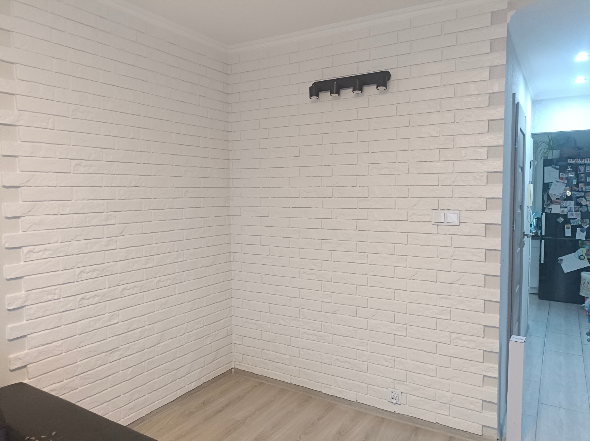 Wykończony róg pokoju z białą ścianą imitującą cegłę, oświetleniem punktowym i panelami podłogowymi. Widok na wnętrze pomieszczenia przez otwarte drzwi.