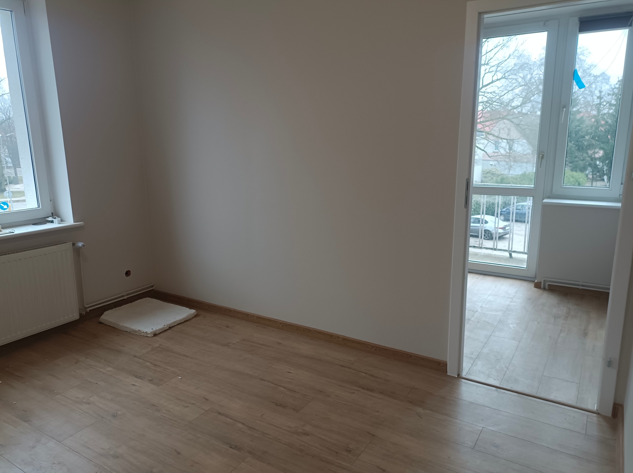 Jasne wnętrze z drewnianą podłogą, białymi ścianami i grzejnikiem pod oknem. Widok na balkon z przeszklonymi drzwiami. Fragment płyty przy grzejniku.