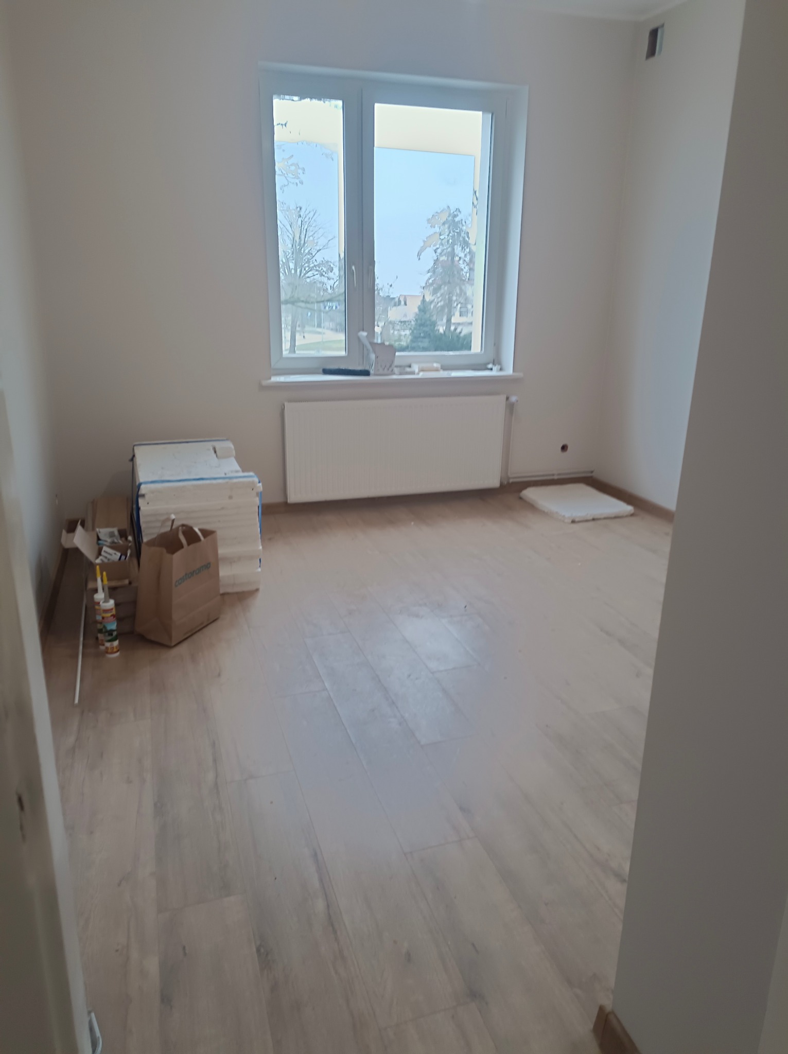 Jasne panele podłogowe w pokoju z oknem. Przy ścianie materiały budowlane i torba Castorama. Widok na zewnątrz przez okno.