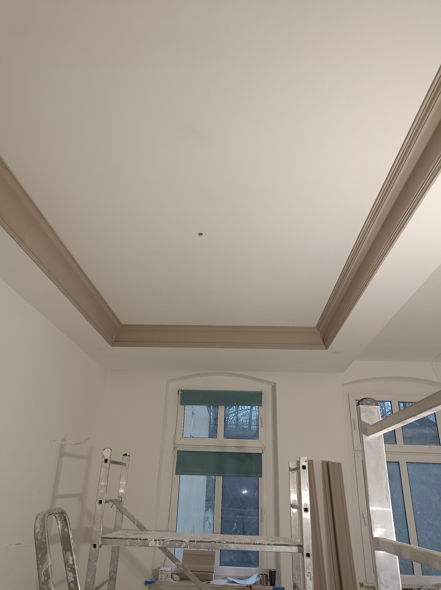 Wykończony sufit z dekoracyjną listwą w odcieniach beżu, widoczny z perspektywy podłogi. W tle okna i drabiny malarskie, sugerujące prace wykończeniowe.