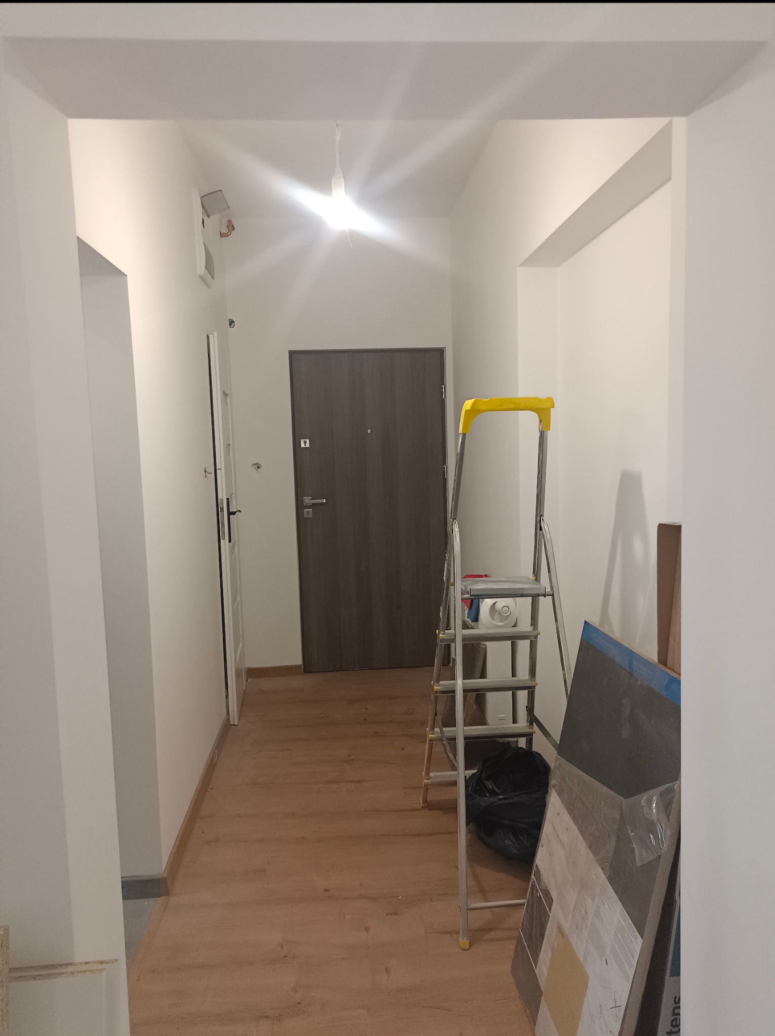 Korytarz w trakcie prac wykończeniowych: drabina, folia malarska, panele podłogowe i świeżo pomalowane ściany na biało. Widoczne drzwi wejściowe i ościeżnica.