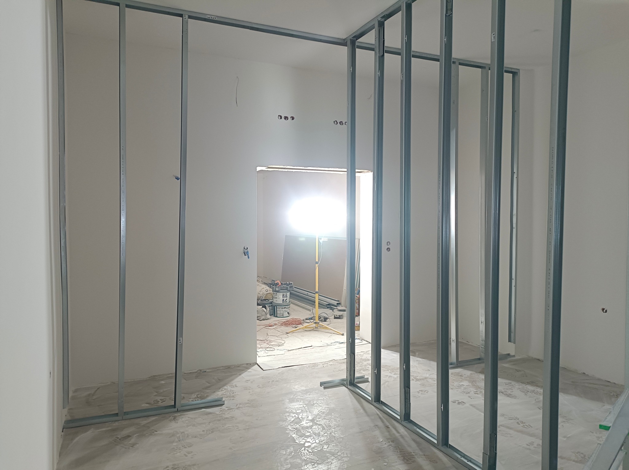 Metalowy szkielet ściany działowej na podłodze, widoczne przewody elektryczne w ścianie, przygotowanie do montażu płyt. W tle narzędzia i lampa oświetleniowa.