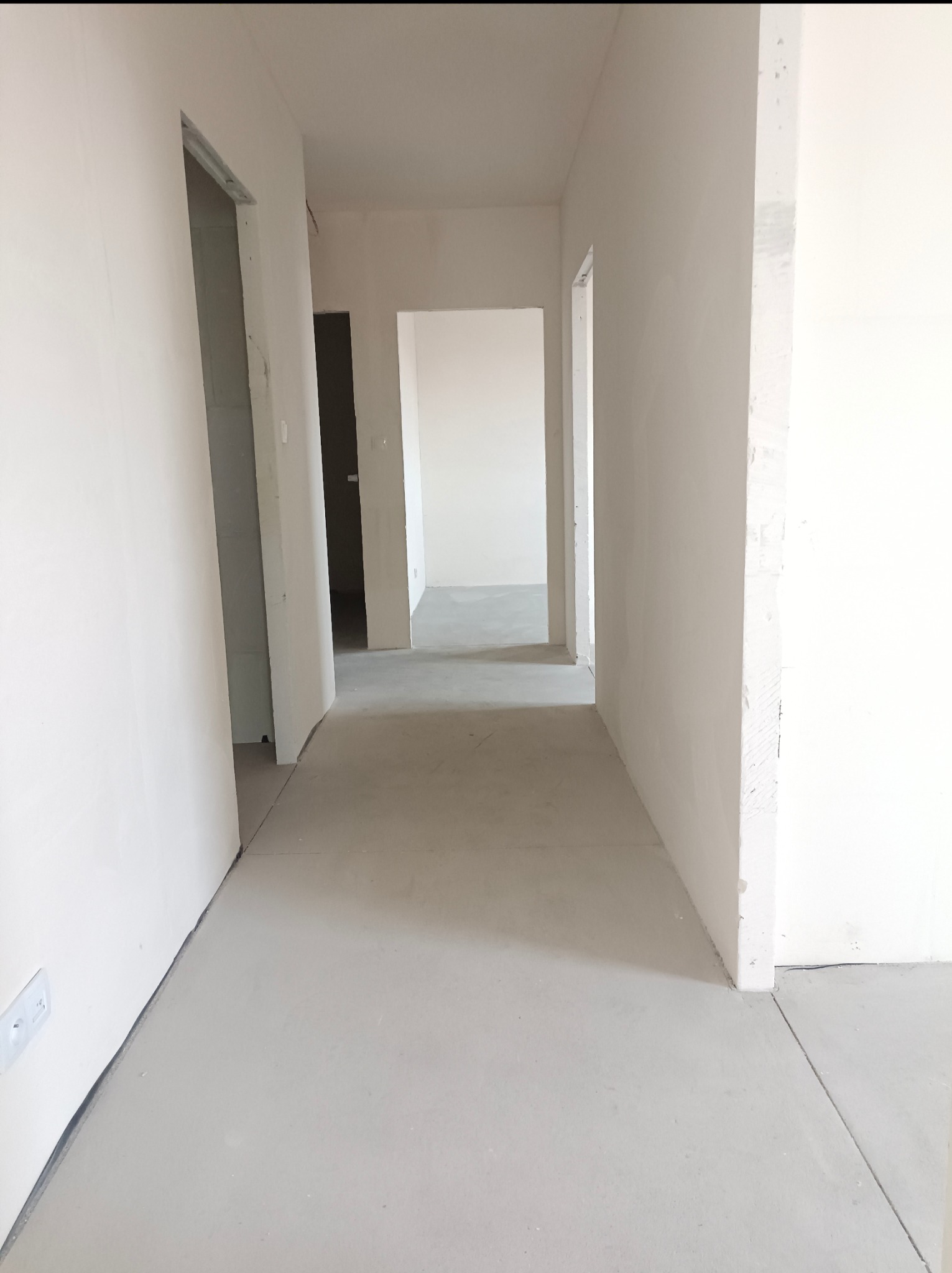 Surowe wnętrze w trakcie prac wykończeniowych. Widok na korytarz z otwartymi przejściami, ściany przygotowane do malowania, wylewka podłogowa. Minimalistyczny, jasny kadr.