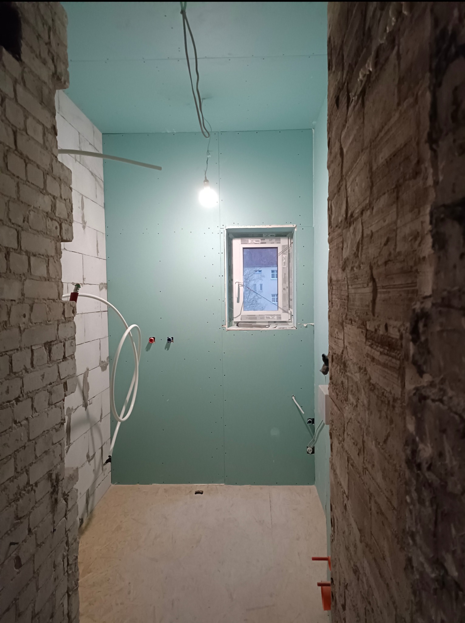 Wnętrze w trakcie prac: ściany z surowej cegły kontrastują z płytami gipsowymi, widoczne instalacje wodne i elektryczne, okno z widokiem na zewnątrz.