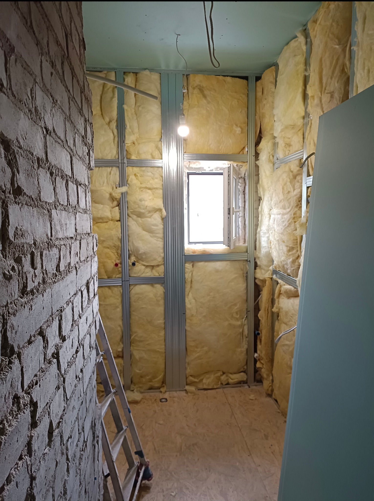 Ocieplanie ścian wełną mineralną, widoczny stelaż z profili stalowych i fragment ściany z cegły. W tle okno. Prace wykończeniowe wewnątrz budynku.