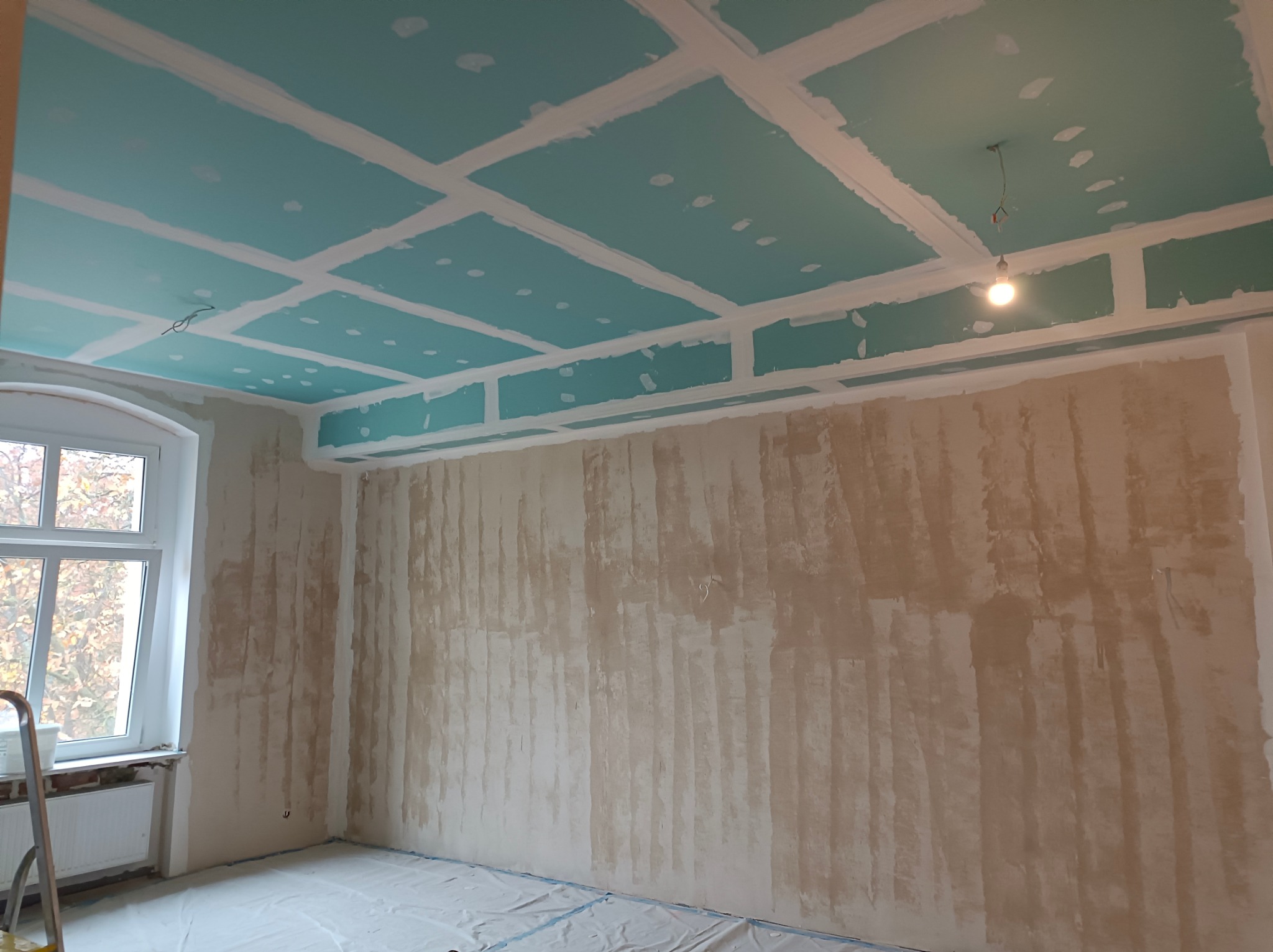 Wnętrze w trakcie prac: sufit z płyt g-k w kolorze turkusowym, białe listwy, surowe ściany przygotowane do malowania, okno z widokiem na drzewa, podłoga zabezpieczona folią.
