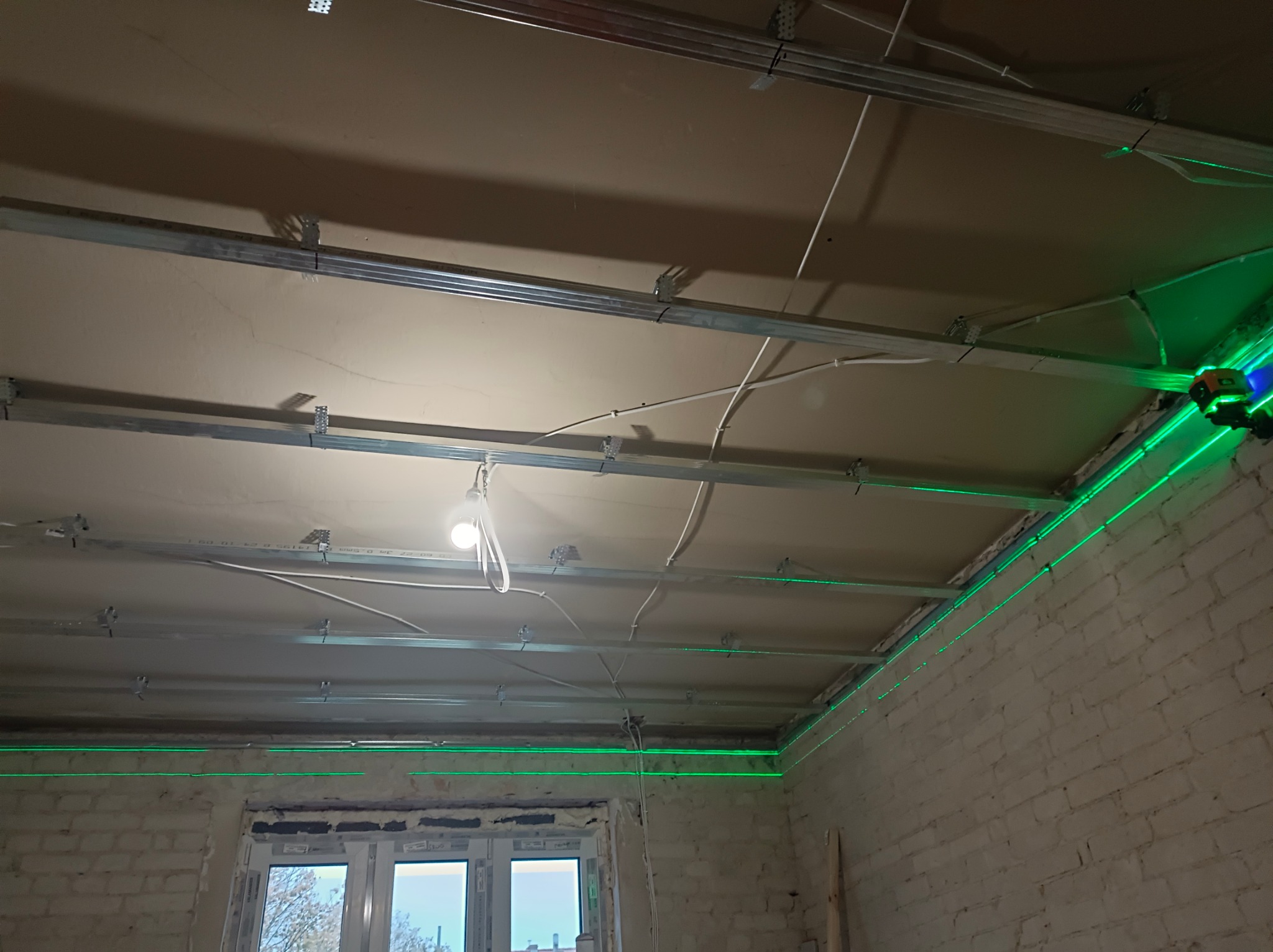 Montaż sufitu podwieszanego z profili, wyznaczanie poziomu laserem krzyżowym, widoczne przewody elektryczne i ceglana ściana. Etap prac wykończeniowych.