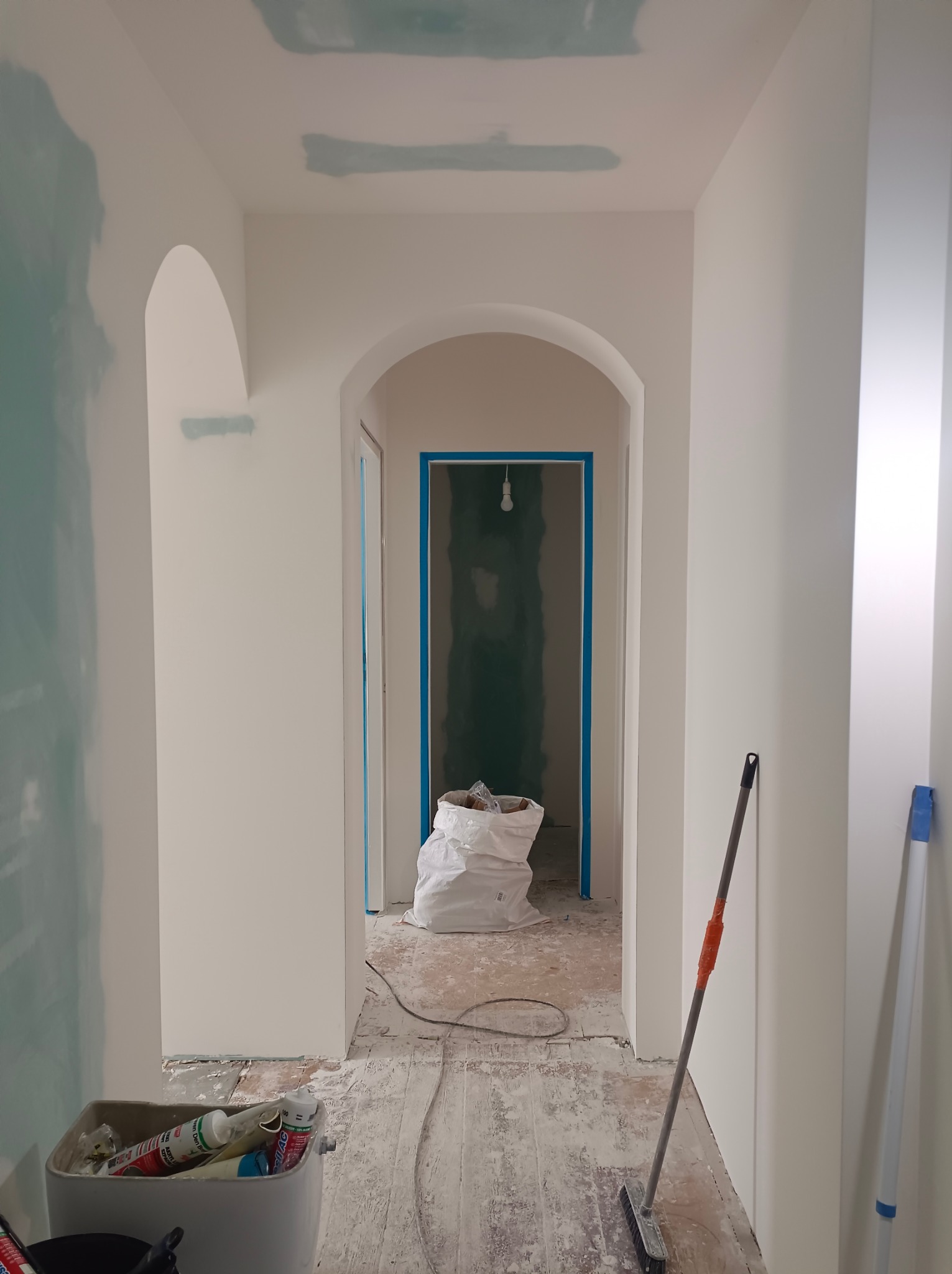 Korytarz w trakcie malowania: białe ściany z łukowymi przejściami, widoczne ślady po szpachlowaniu, narzędzia i worek z materiałami budowlanymi na podłodze.