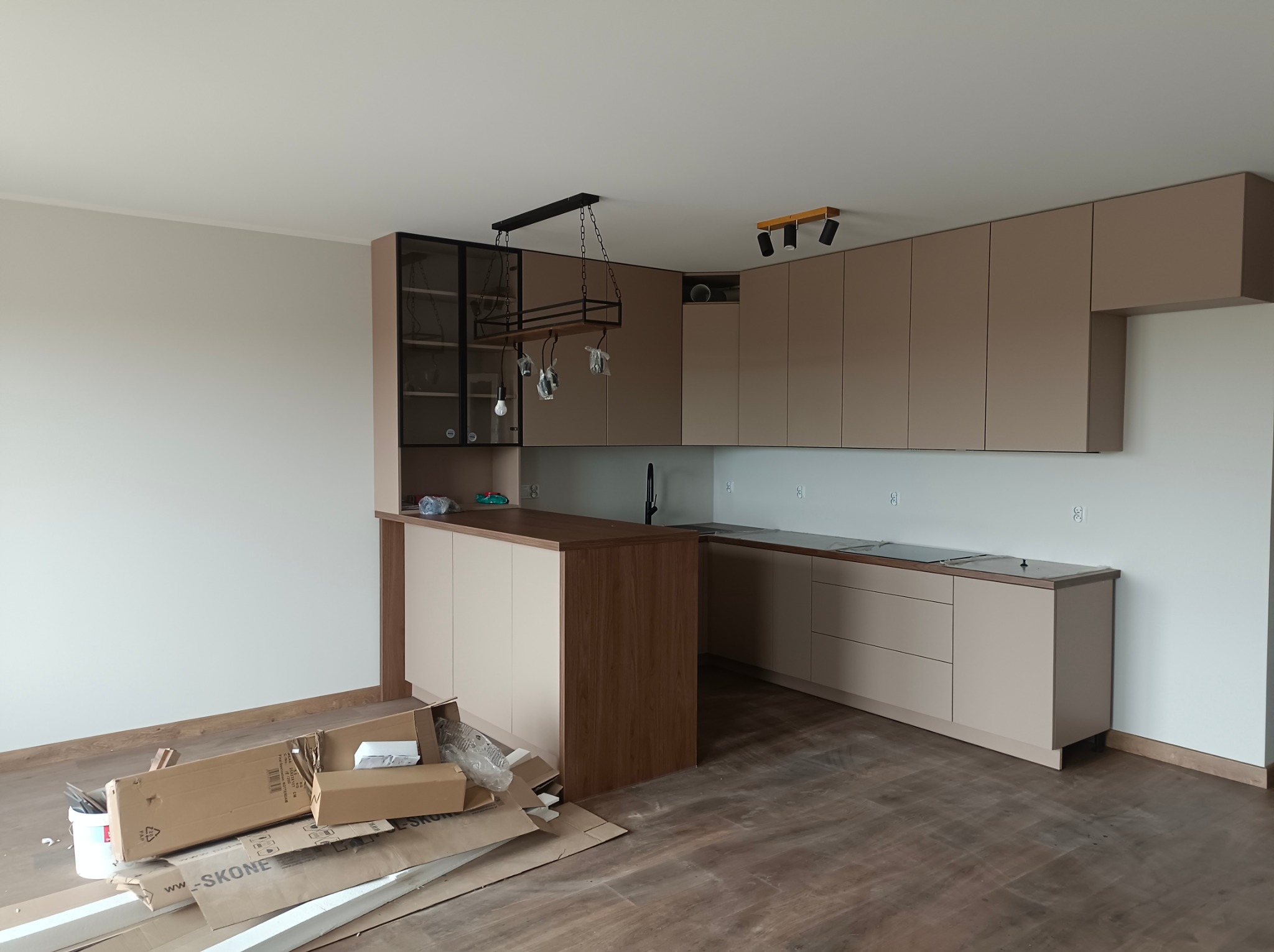 Beżowa zabudowa kuchenna z wyspą i oświetleniem w surowym wnętrzu. Na podłodze kartony i ślady po remoncie. Nowoczesny design.