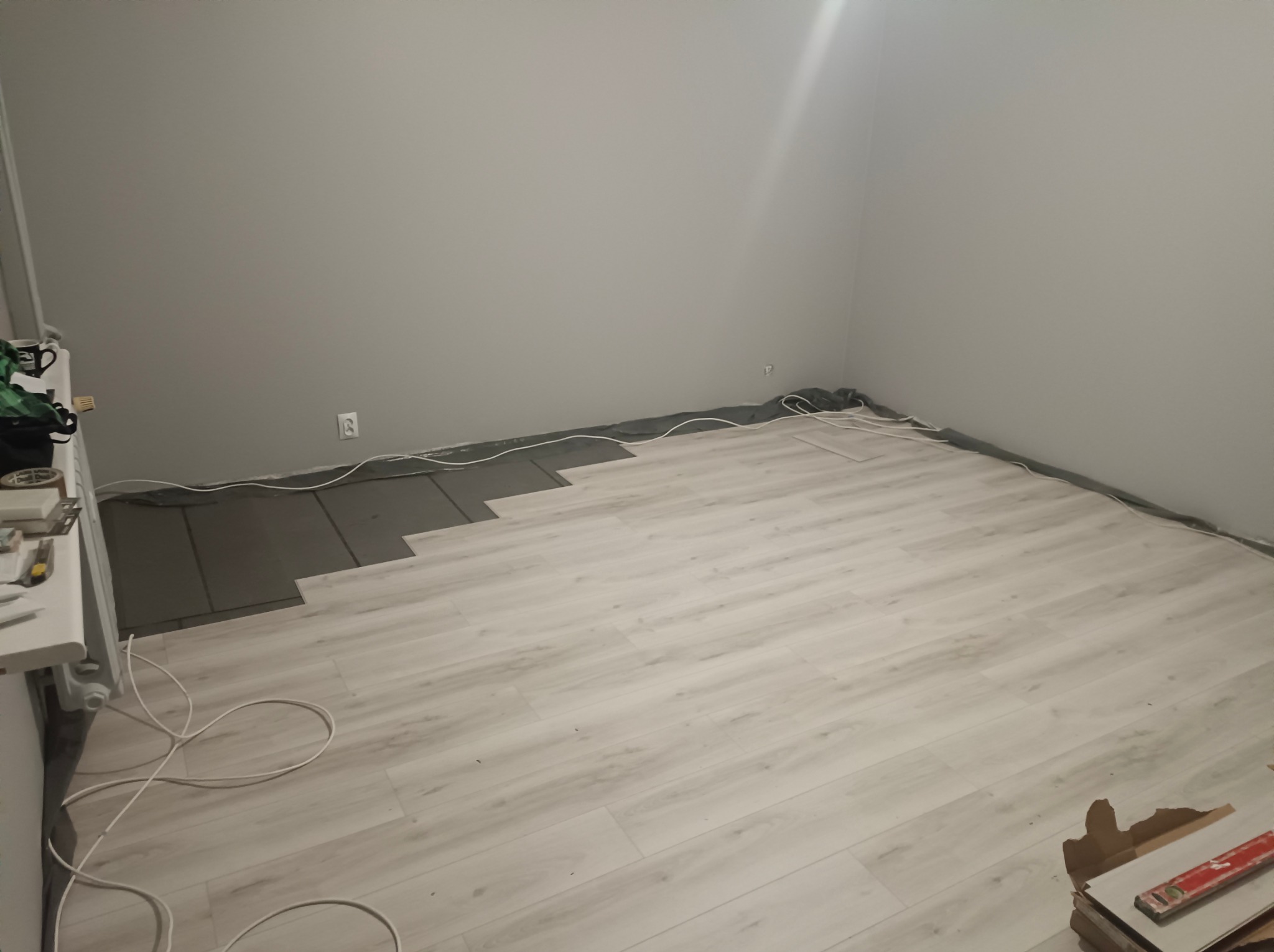 Fragment układania podłogi z paneli w jasnym odcieniu drewna. Widoczna część podkładu i narzędzia. Ściany szare, surowe, z widocznym gniazdkiem elektrycznym.