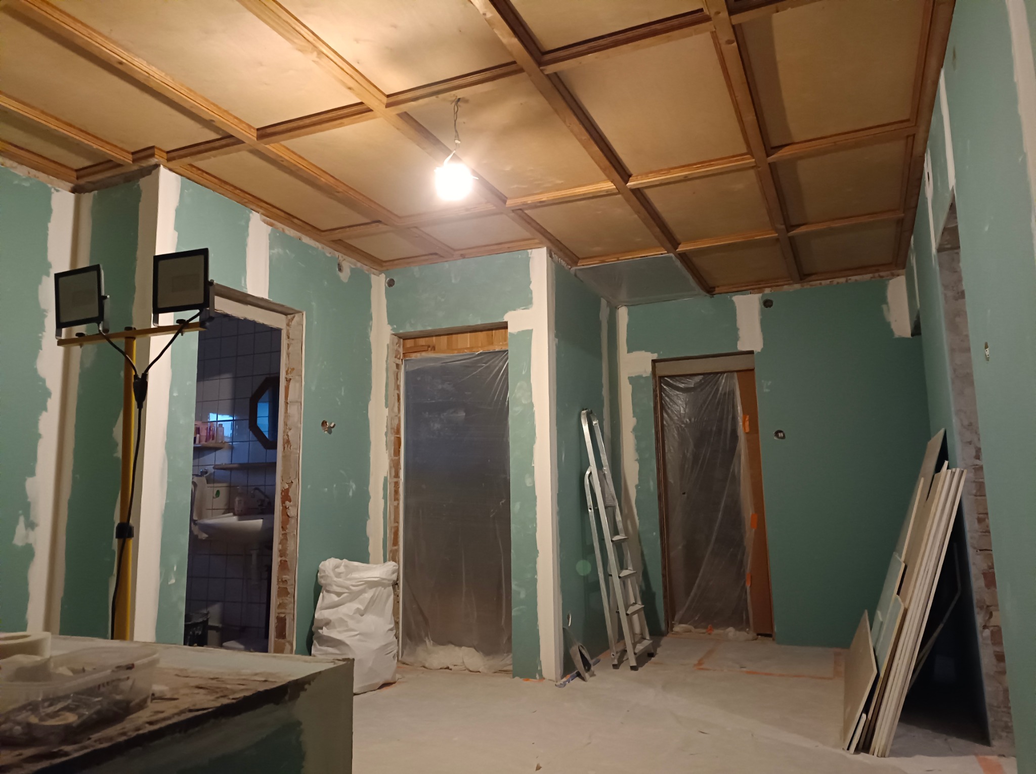 Remont wnętrza: ściany z zieloną farbą i gładzią, drewniany sufit kasetonowy, drabina, płyty gipsowe. Widoczne przygotowania do wykończenia.