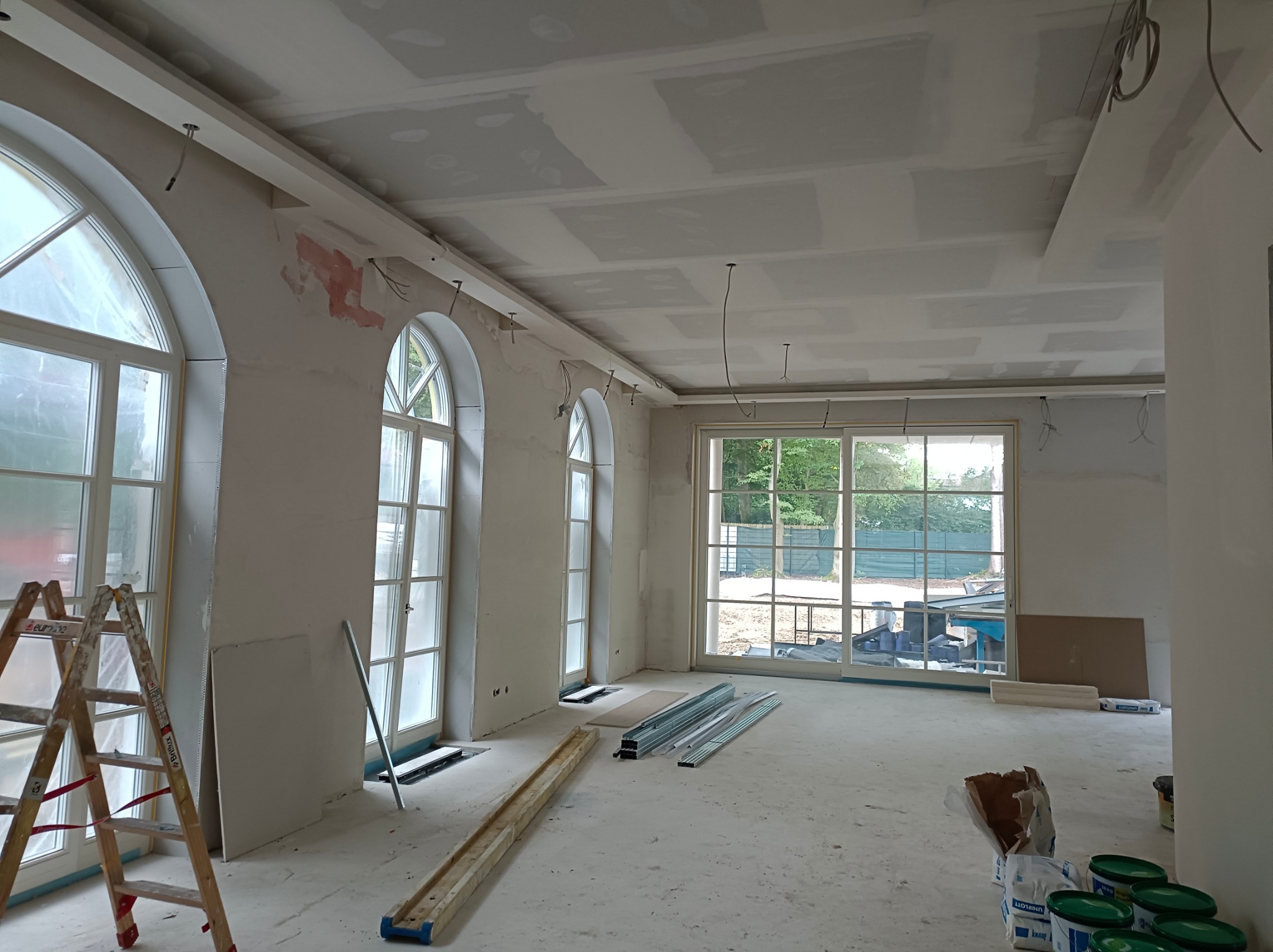 Wnętrze w trakcie prac: widoczne płyty gipsowe na suficie, ściany przygotowane do malowania, okna łukowe. Na podłodze materiały budowlane i drabina.