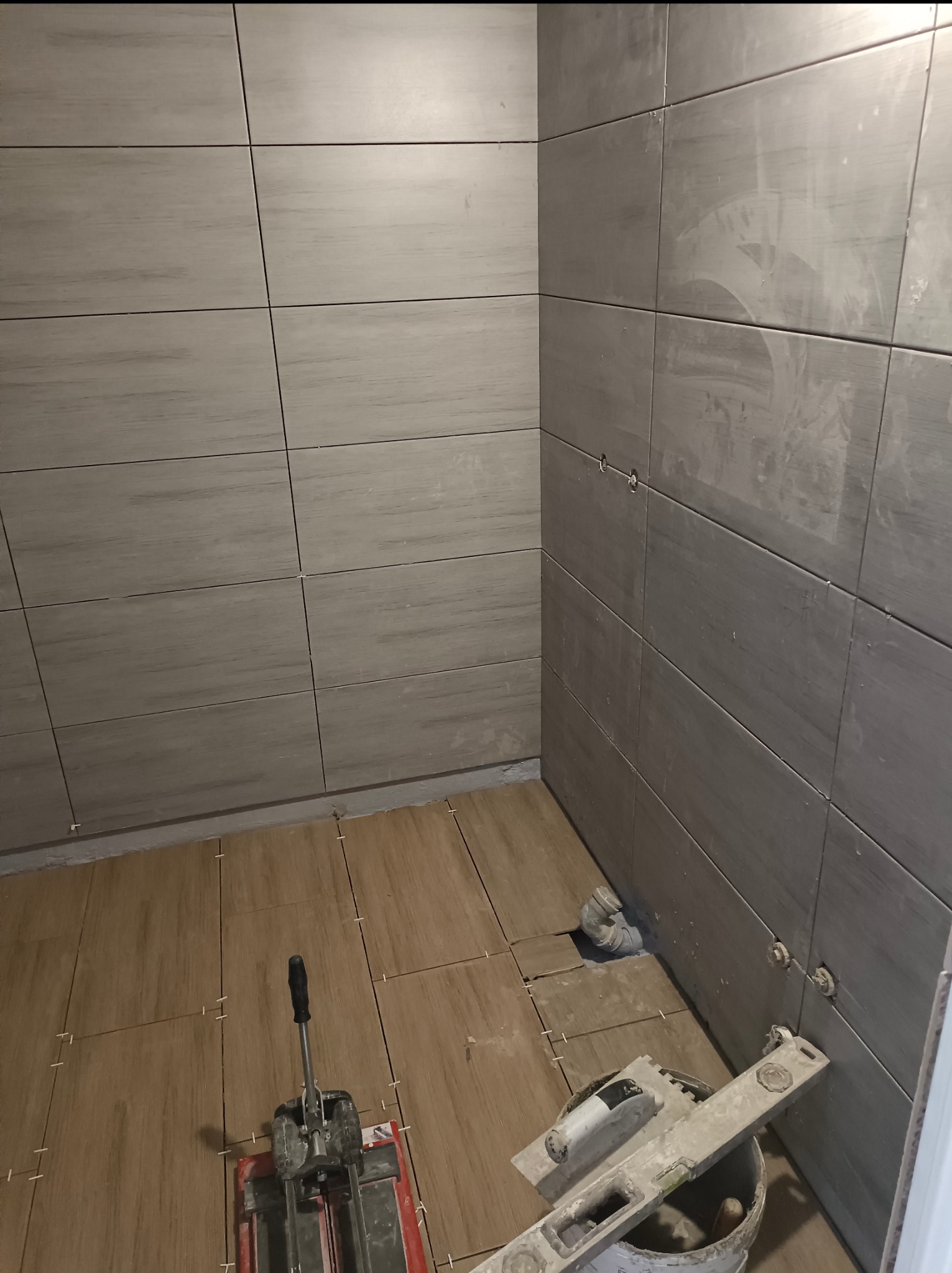 Układanie płytek drewnopodobnych na podłodze i szarych na ścianach w łazience. Widoczny sprzęt glazurniczy i poziomica w wiadrze z zaprawą.