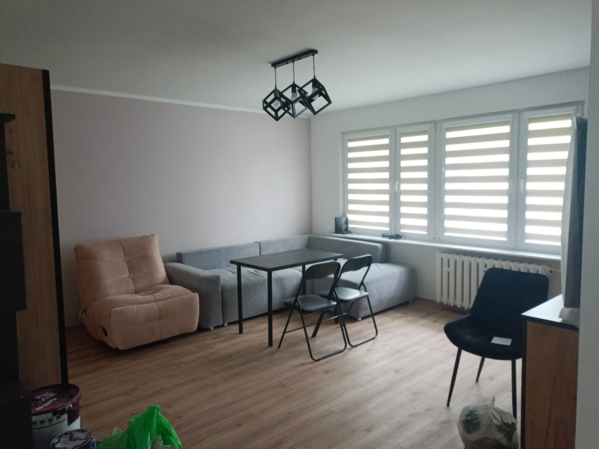 Jasne wnętrze pokoju dziennego z szarą sofą, stolikiem i krzesłami na drewnianej podłodze. Okno z żaluzjami i nowoczesna lampa sufitowa.