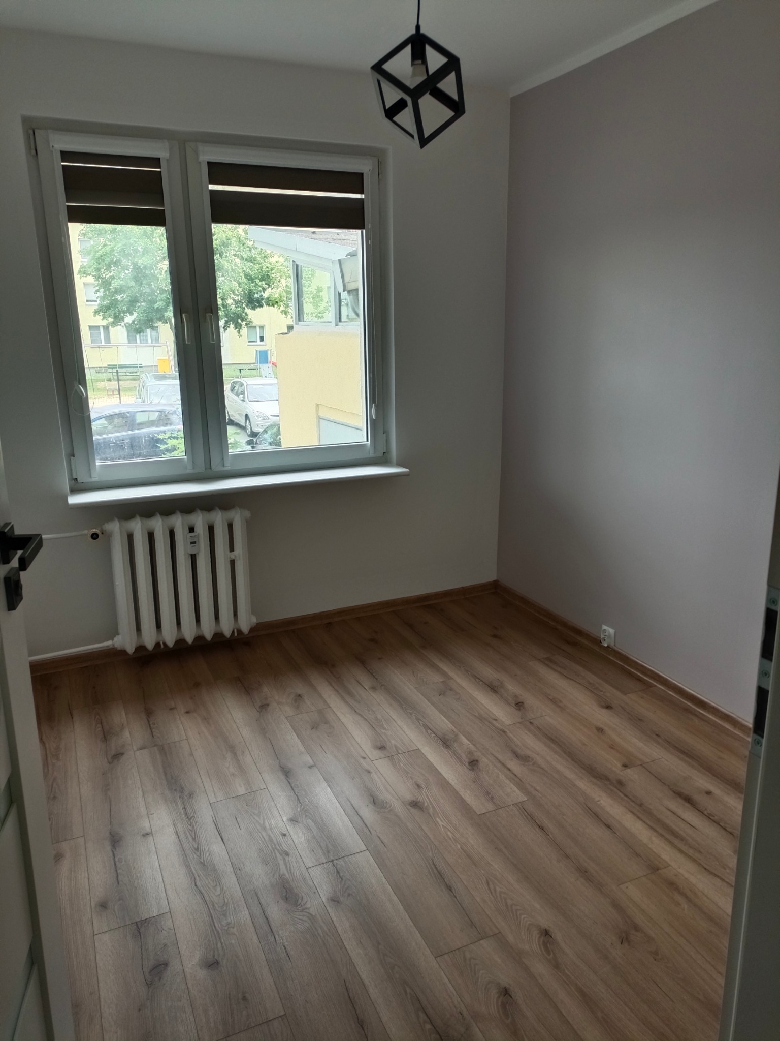 Pusty pokój po remoncie: drewniana podłoga, grzejnik pod oknem z roletami, nowoczesna lampa sufitowa. Ściany pomalowane na jasny odcień szarości.