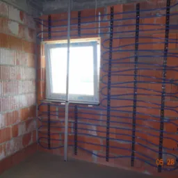 Ściana z cegły z zamontowanymi uchwytami i niebieskimi rurami, prawdopodobnie instalacja ogrzewania podłogowego. Widoczne okno i betonowa podłoga w surowym stanie.