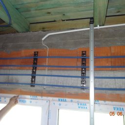 EKO-ENERGIA ZAKŁAD PRODUKCYJNO USŁUGOWO HANDLOWY Sławomir Wajs - Wnętrze w trakcie prac: niebieskie rury przymocowane do ściany za pomocą czarnych uchwytów, biała rurka biegnąca pod sufitem, miarka pionowo przy rurach.