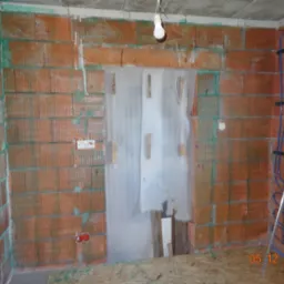 Ceglana ściana w trakcie budowy z widocznymi przewodami elektrycznymi i instalacją ogrzewania podłogowego. Na ścianie gniazdka elektryczne i wisząca żarówka.