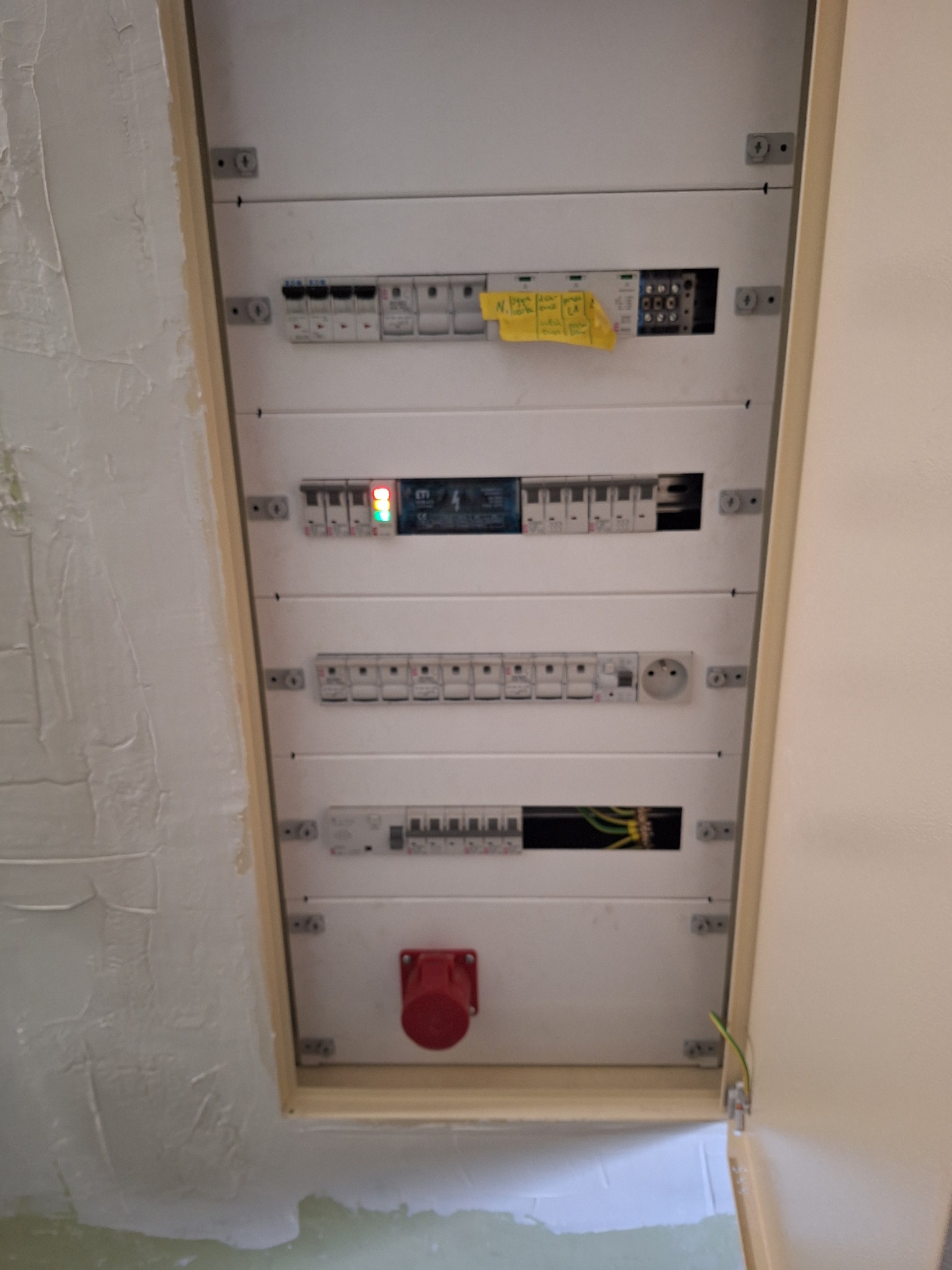 Biała skrzynka rozdzielcza z bezpiecznikami i gniazdem siłowym na ścianie z surowym tynkiem. Widoczne przewody i elementy instalacji elektrycznej.