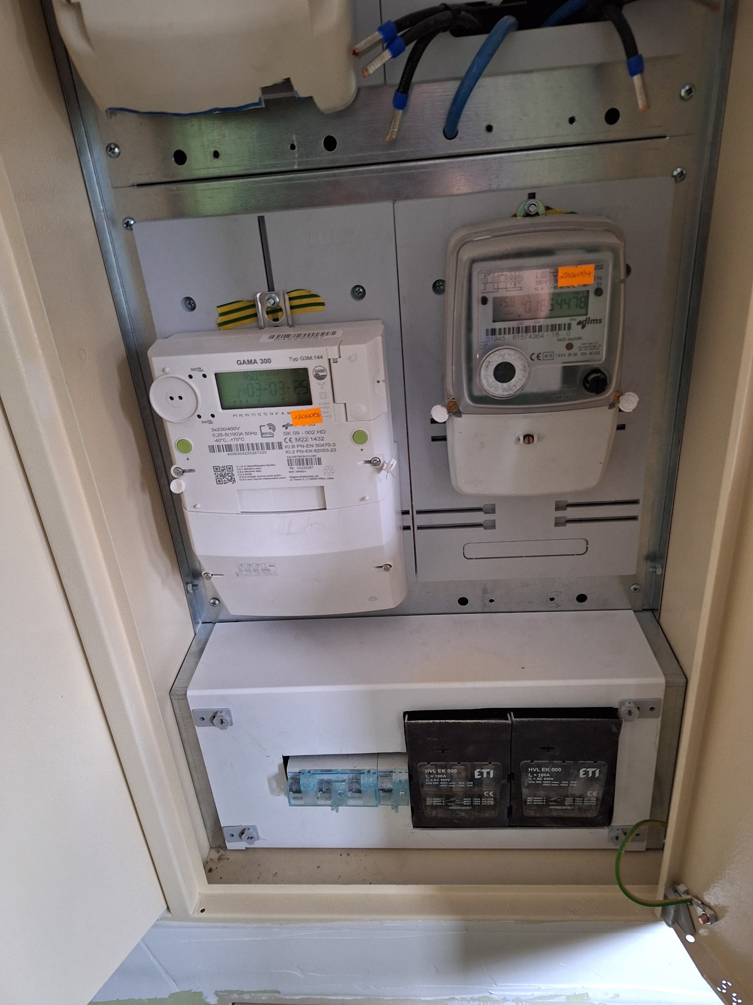 Rozdzielnia elektryczna z dwoma licznikami (cyfrowym i analogowym) oraz zabezpieczeniami, widoczne przewody i elementy montażowe.