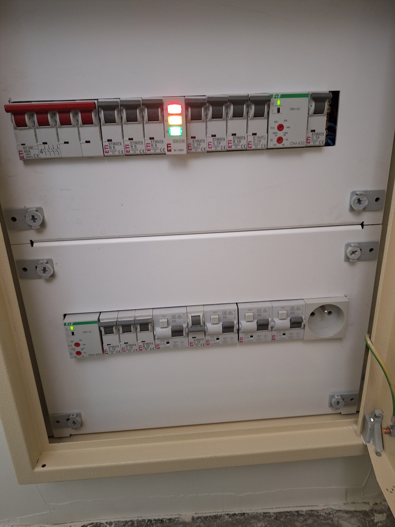 Rozdzielnica elektryczna z bezpiecznikami, niektóre włączone (świecące diody), widoczna instalacja i gniazdko. Obudowa biała, mocowana na śruby.