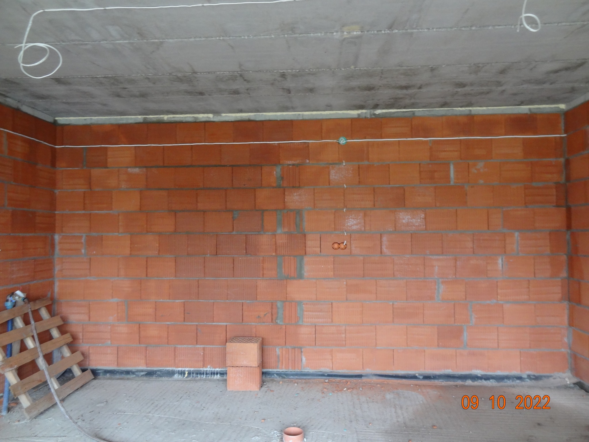 Czerwona ściana z pustaków ceramicznych w surowym stanie, z widocznymi przewodami elektrycznymi u góry i na suficie, w trakcie budowy.