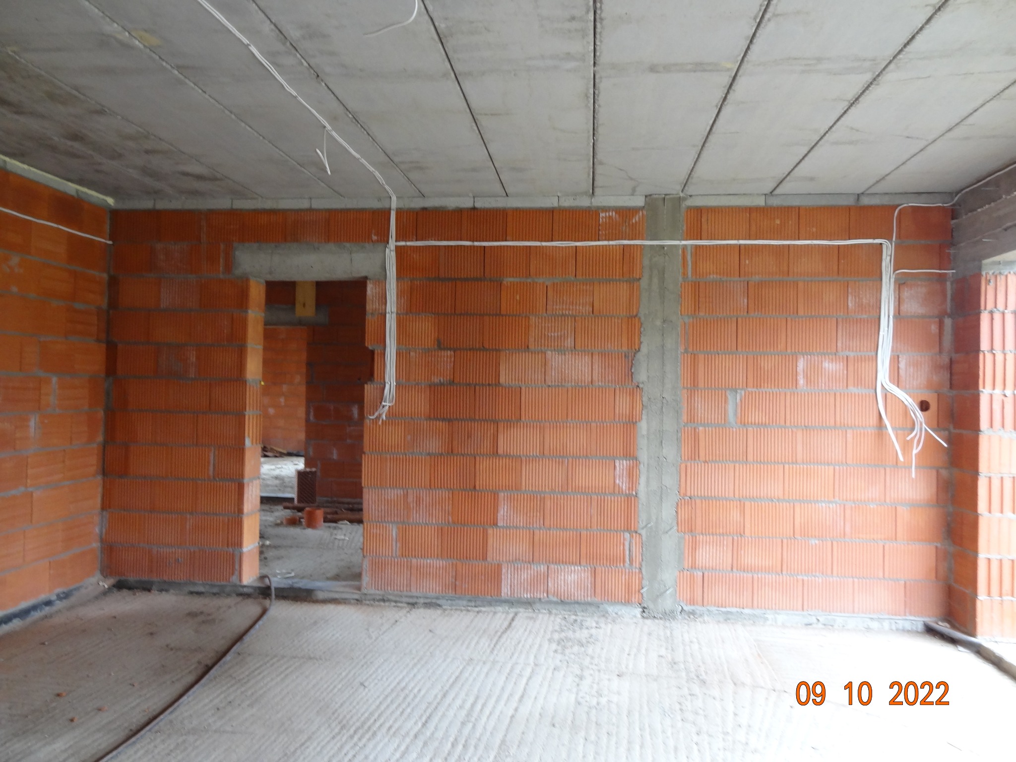 Wnętrze w stanie surowym: ściany z czerwonej cegły, betonowy strop z wystającymi kablami elektrycznymi. Widoczne przewody i otwory instalacyjne.