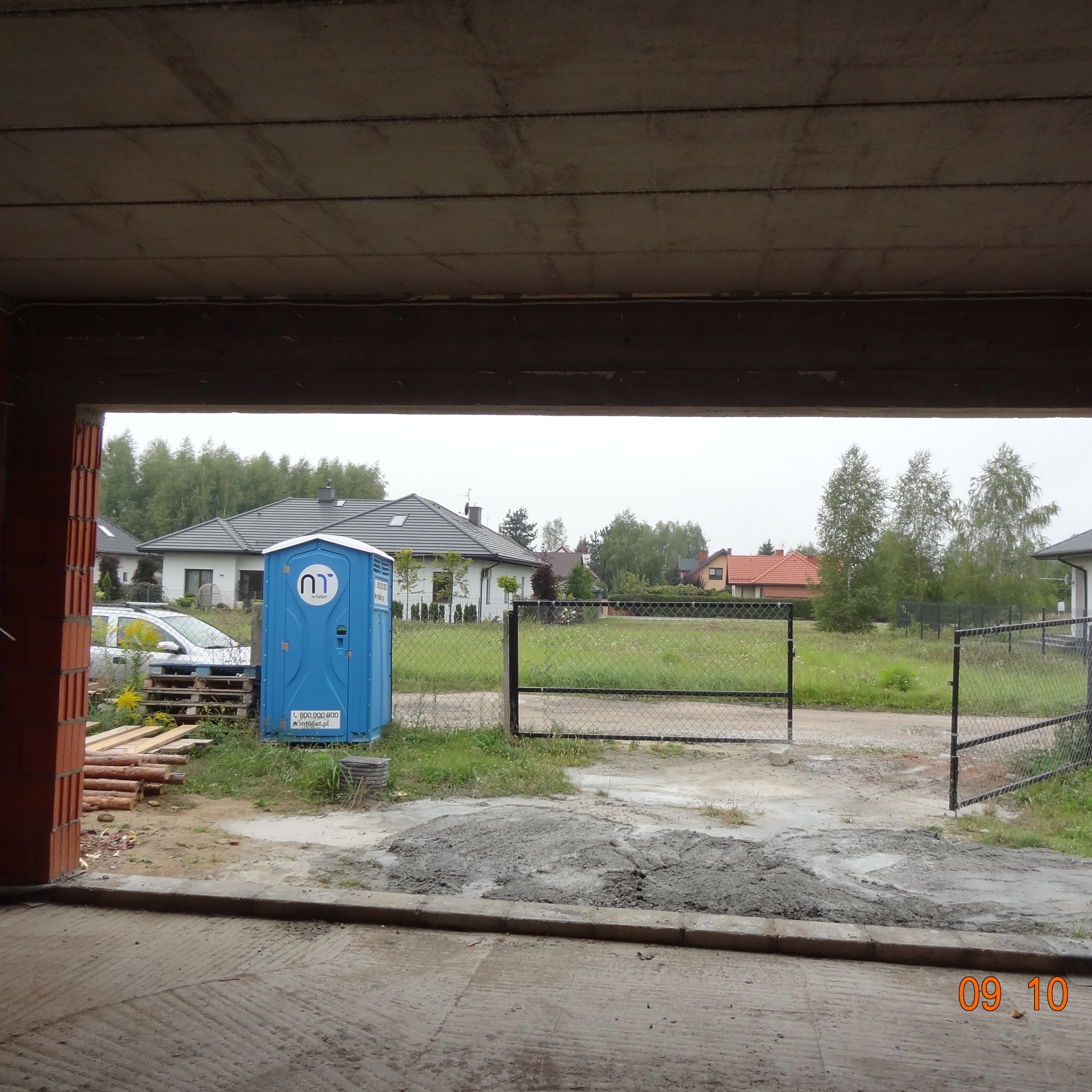 Widok z wnętrza budynku w trakcie budowy na podwórze z przenośną toaletą, ogrodzeniem i domami w tle. Surowy beton na podłodze, cegły i drewno. Dzień pochmurny.
