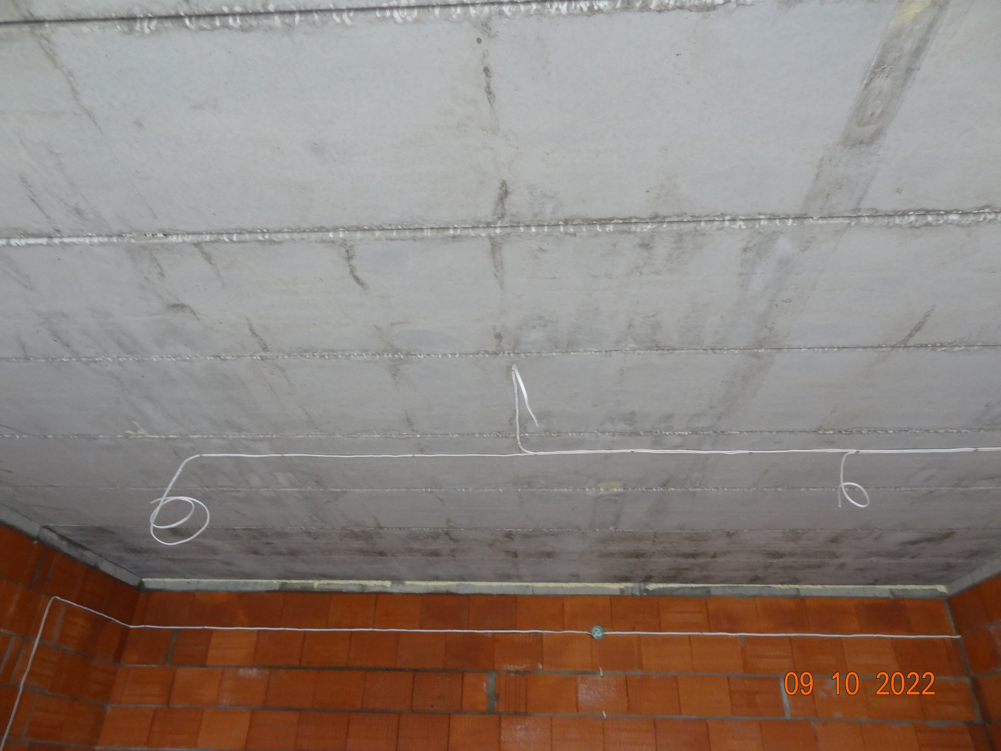 Surowy strop z widocznymi przewodami elektrycznymi, układanymi wzdłuż ceglanej ściany. Kable zwisają luźno, tworząc pętle. Widoczna data zdjęcia w prawym dolnym rogu.