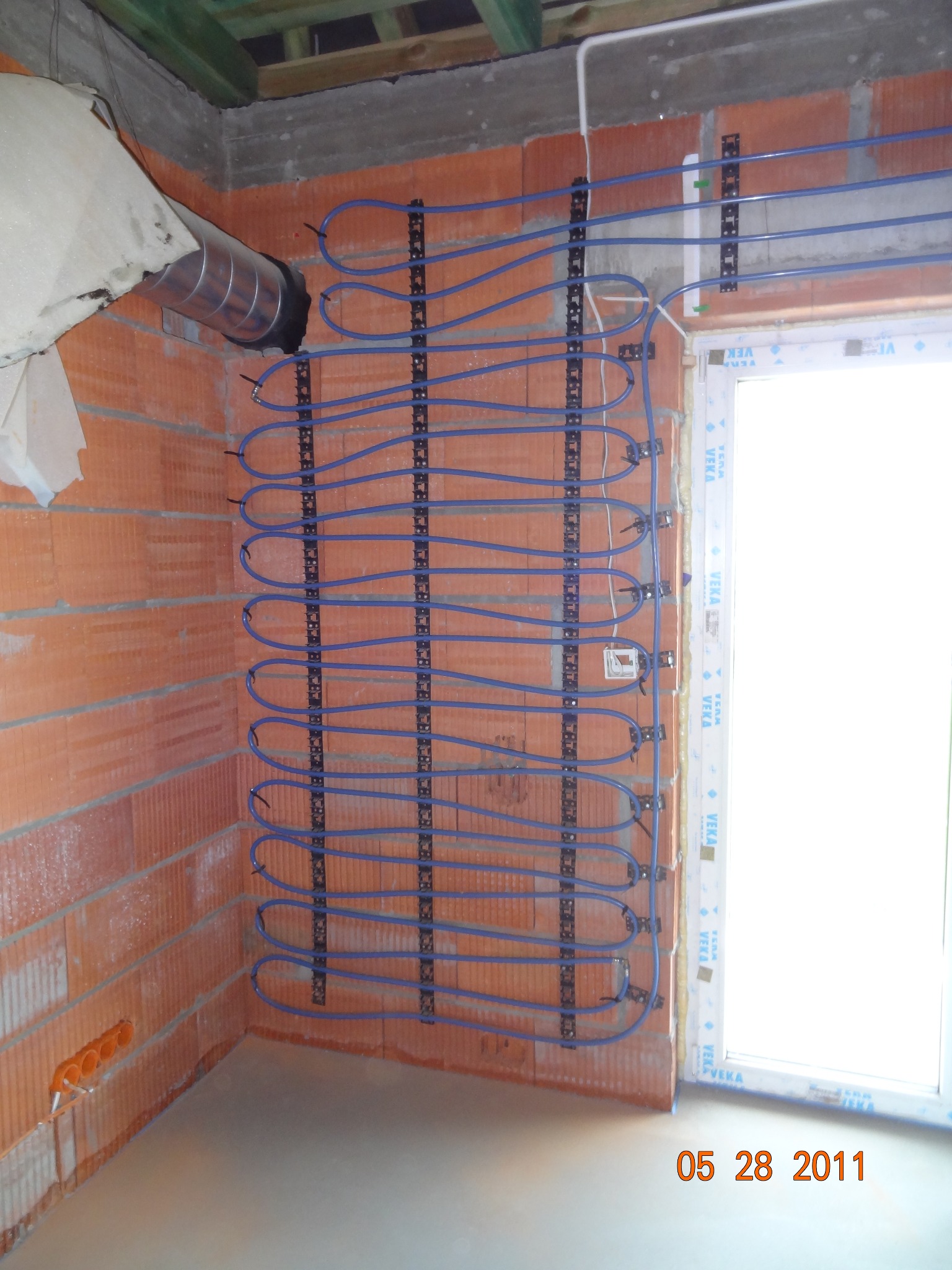 Montaż ogrzewania ściennego z niebieskich rur na ceglanej ścianie, widoczne elementy instalacji wentylacyjnej i elektrycznej, okno w tle. Etap prac wykończeniowych.