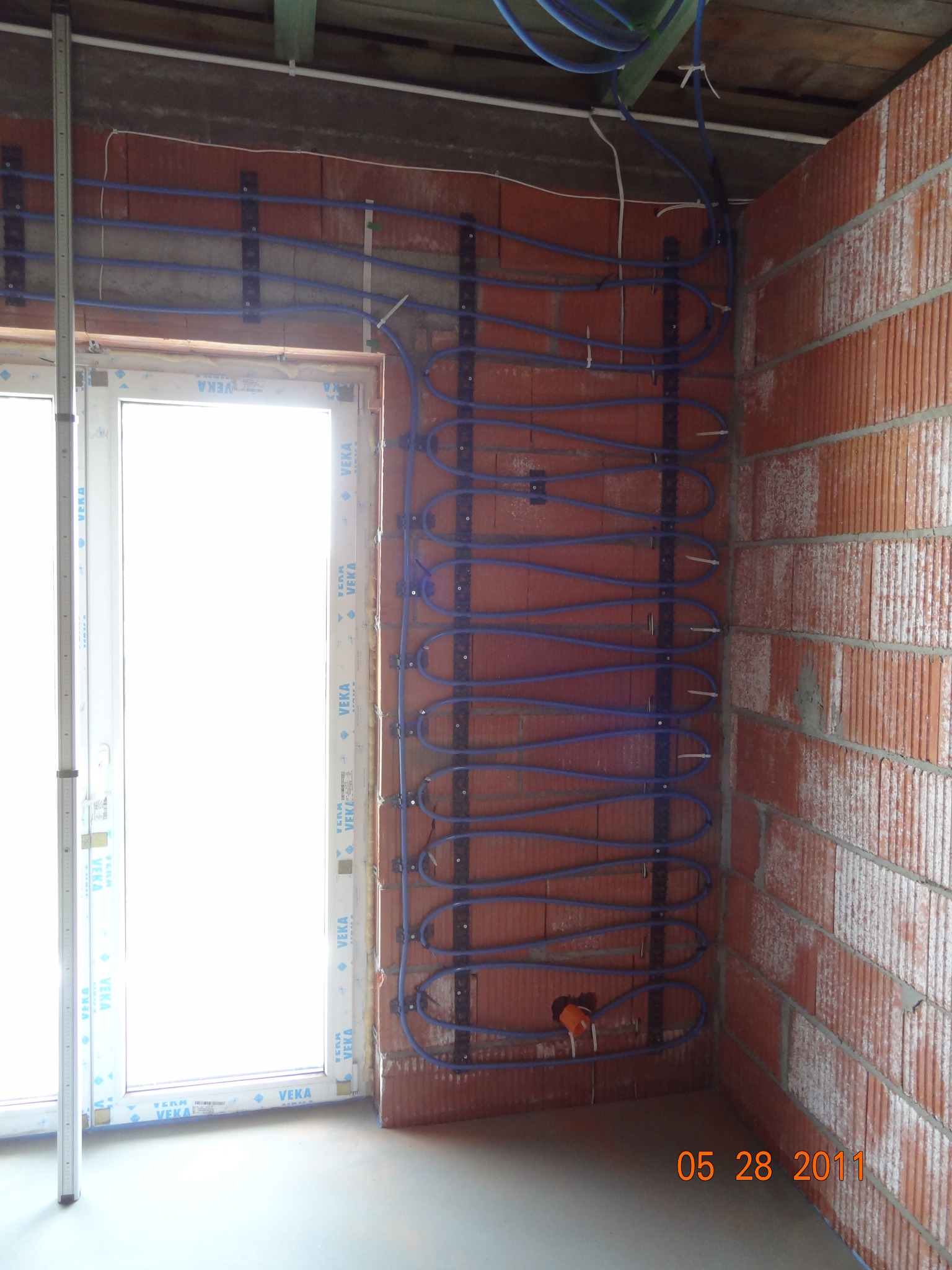 Instalacja ogrzewania podłogowego: niebieskie rury przymocowane do ściany z cegieł, widoczne okno z profilem VEKA, etap prac przed wylaniem posadzki.