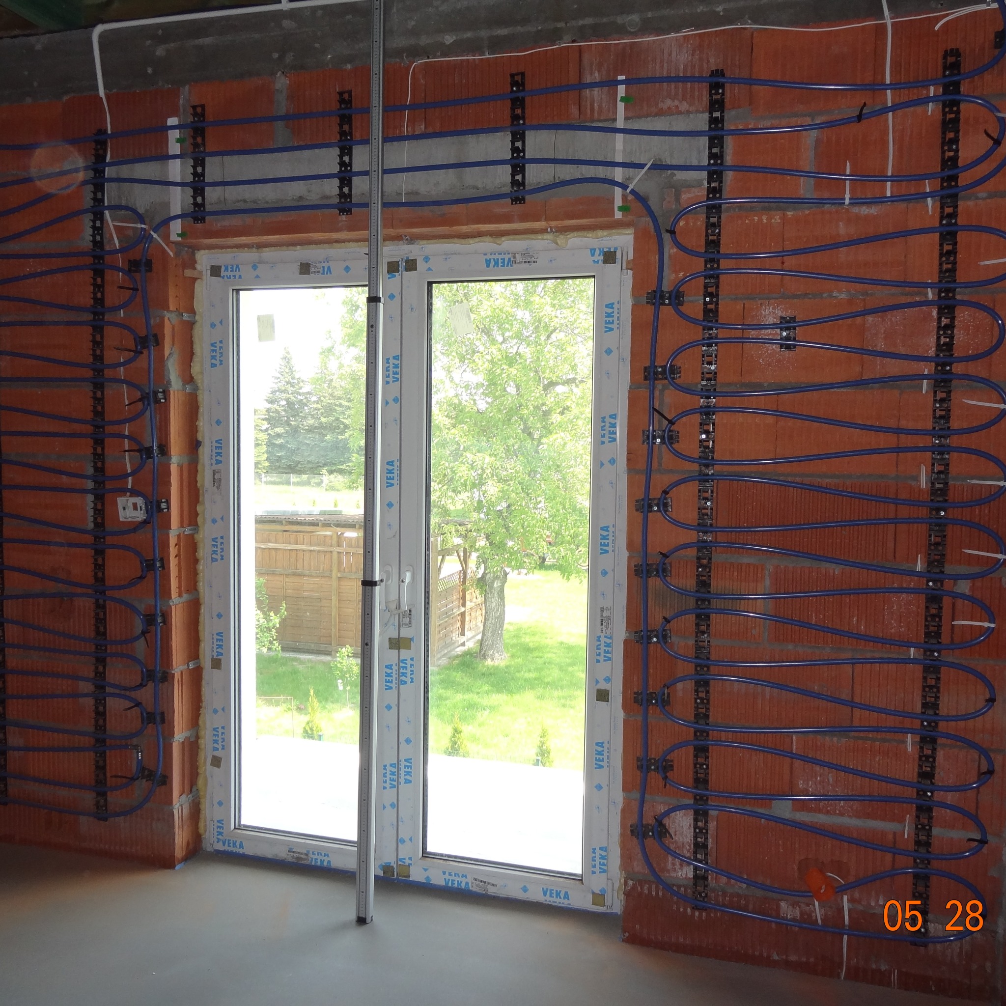 Montaż ogrzewania podłogowego w surowym wnętrzu, z widocznymi niebieskimi rurami przymocowanymi do ceglanych ścian. W tle okno z widokiem na zieleń.