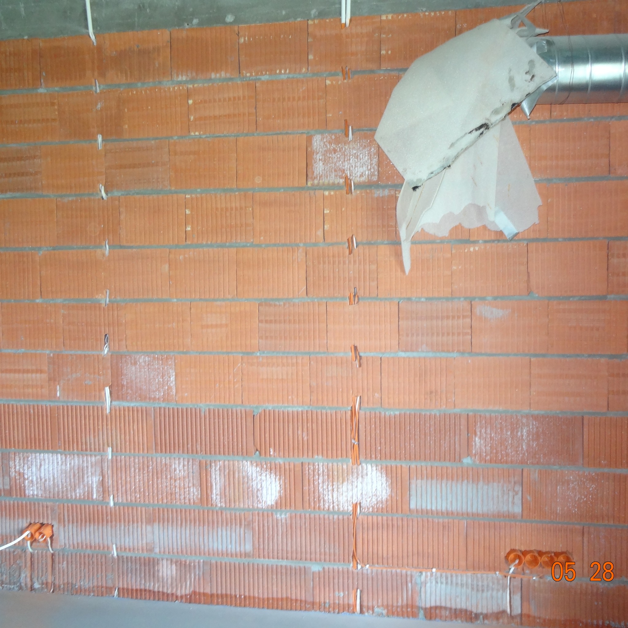 Murowana ściana z widocznymi przewodami elektrycznymi w trakcie montażu, zabezpieczonymi gipsem. Widoczne puszki instalacyjne i fragment wentylacji.