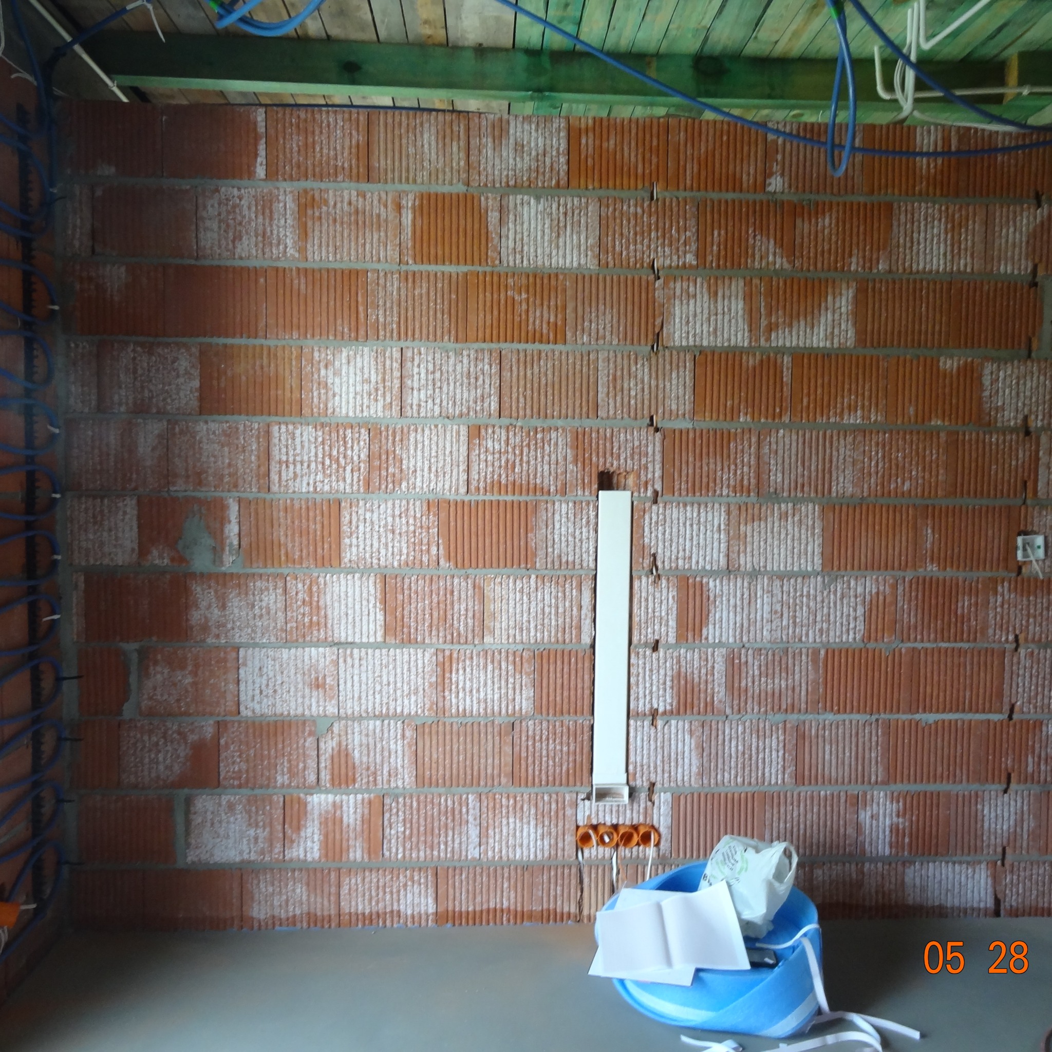 Ściana z cegły dziurawki z widoczną instalacją elektryczną w trakcie montażu, niebieskie kable na ścianie. W tle drewniany strop.