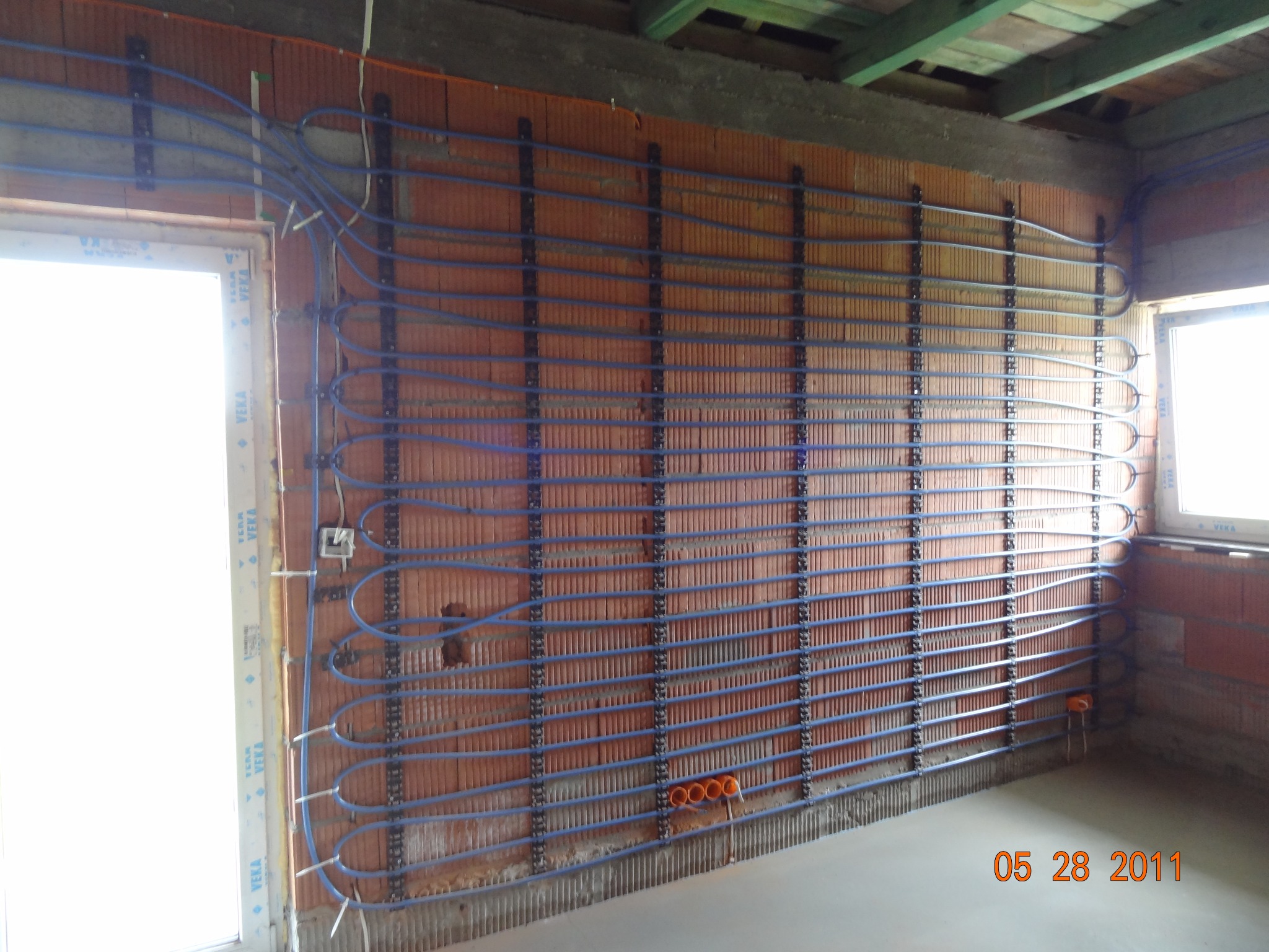 Montaż ogrzewania ściennego: niebieskie rury mocowane do ceglanej ściany na metalowych listwach. Widoczne okno i surowy stan wykończenia wnętrza, instalacja elektryczna.